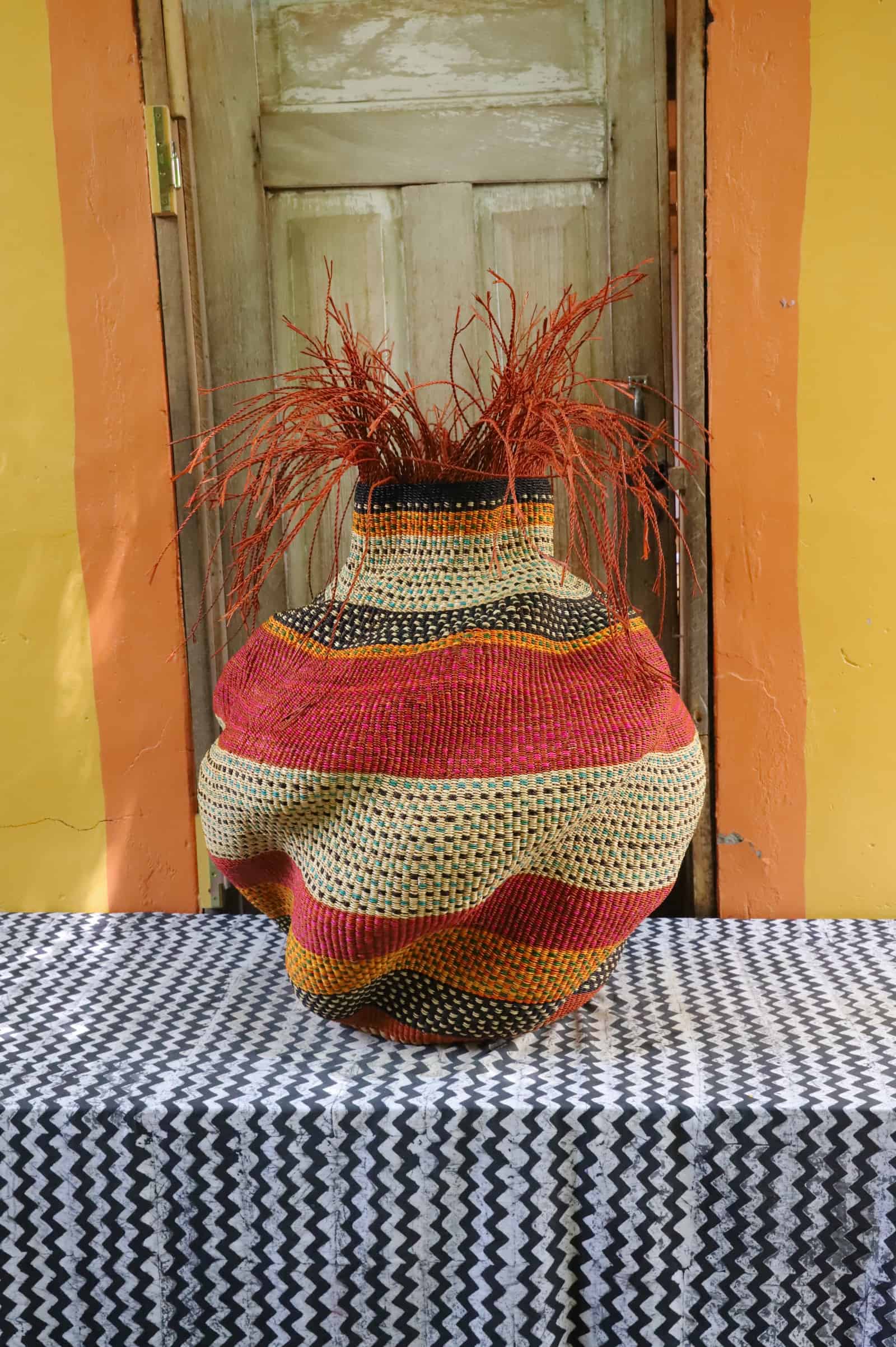 Large Tua-Tia baobab art basket in red and natural stripes with red straw branching, on chevron cloth against a wooden door.