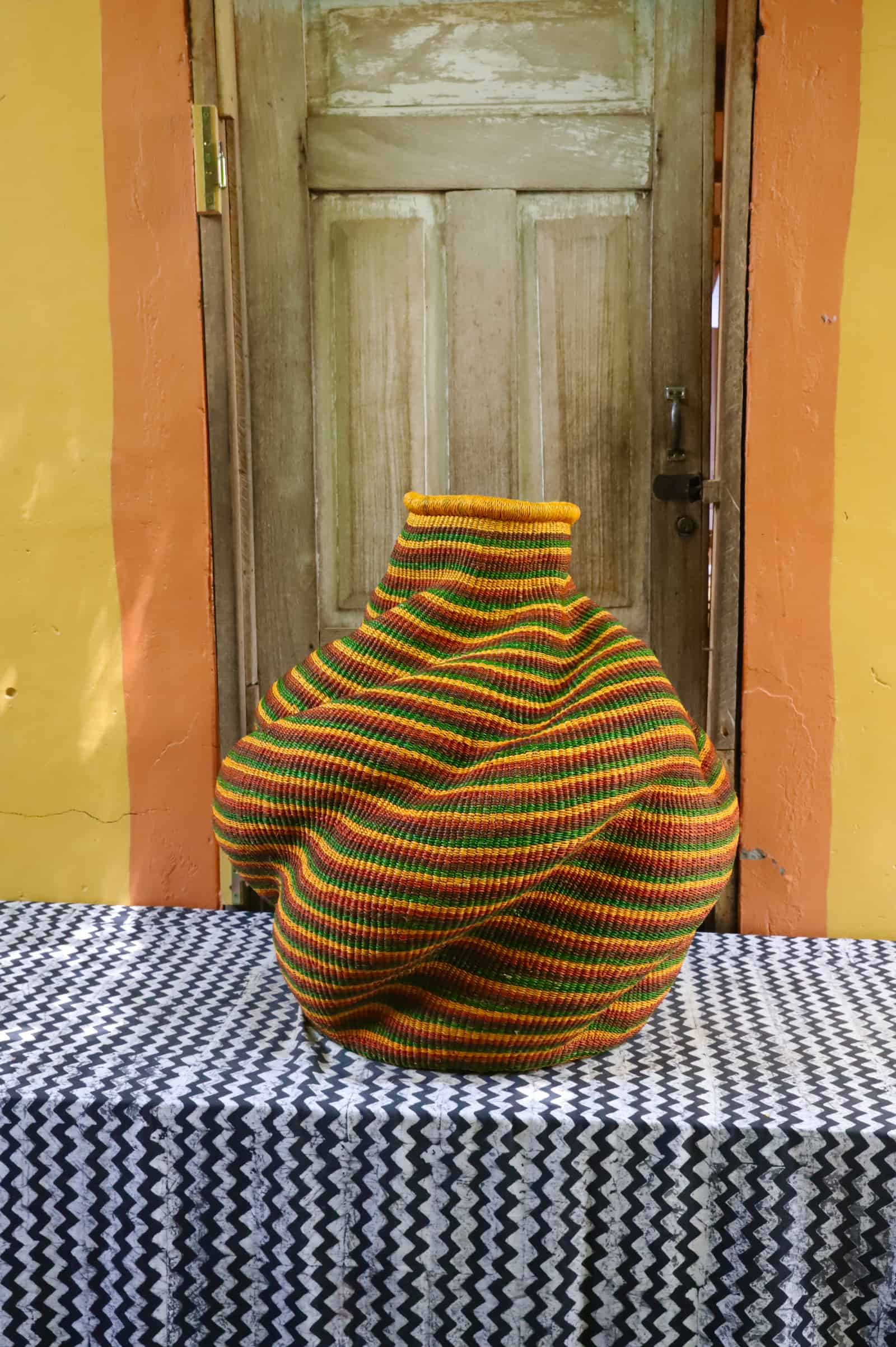 Large handwoven De-engo art basket with a lobed gourd form in amber, green and terracotta stripes, on a chevron cloth.