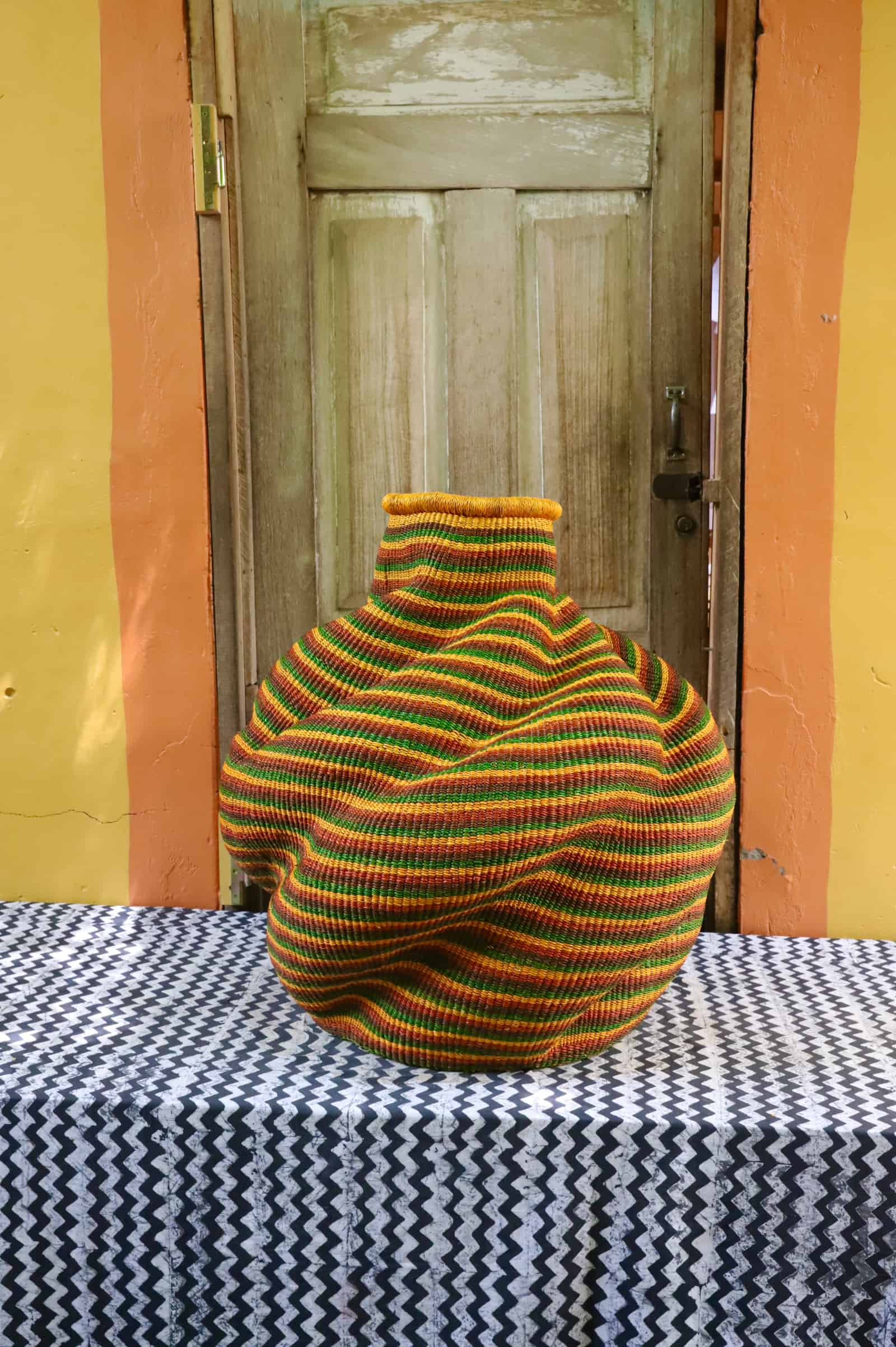 Side view of a large handwoven De-engo art basket in amber, green and terracotta stripes, on a black and white chevron cloth.