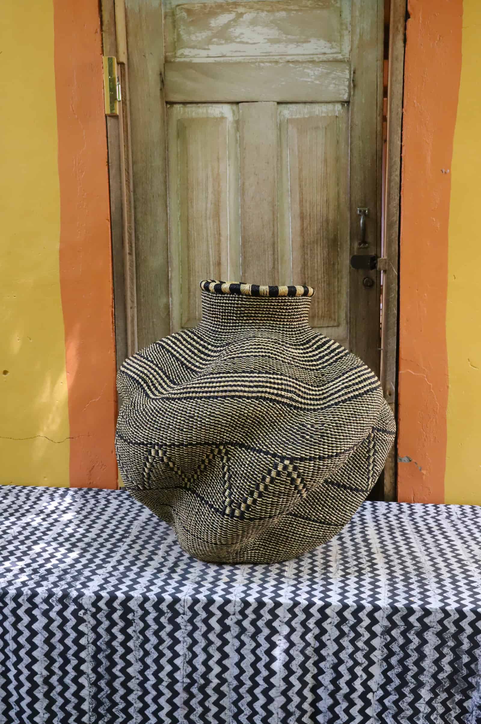 Large handwoven De-engo art basket with a lobed gourd form in natural straw and black geometric pattern, on a chevron cloth.