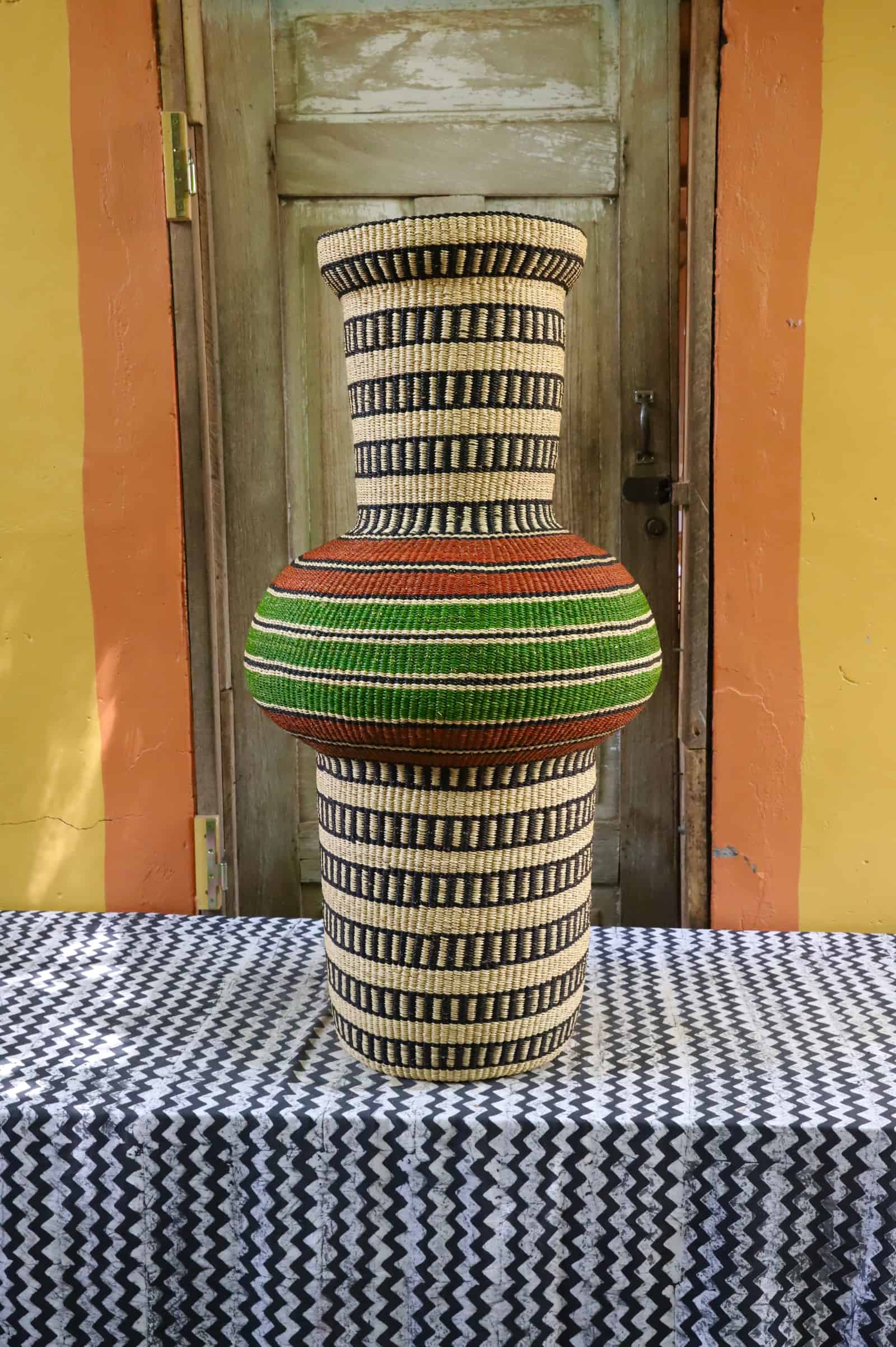 Tall Dugla art basket in natural straw with black horizontal stripes and a green and terracotta band, before a wooden door.