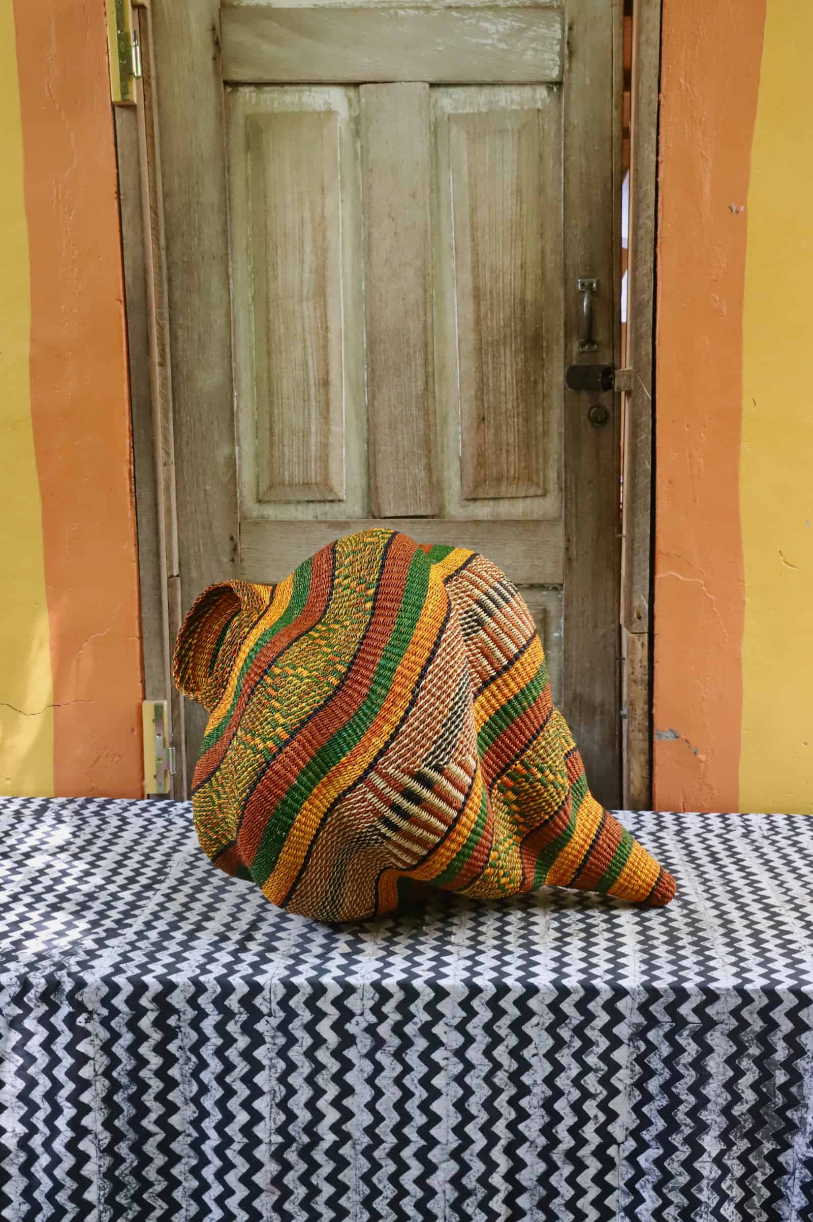 Handwoven Katayeli-tia art basket in amber, terracotta and green flowing stripes, on a black and white chevron cloth.