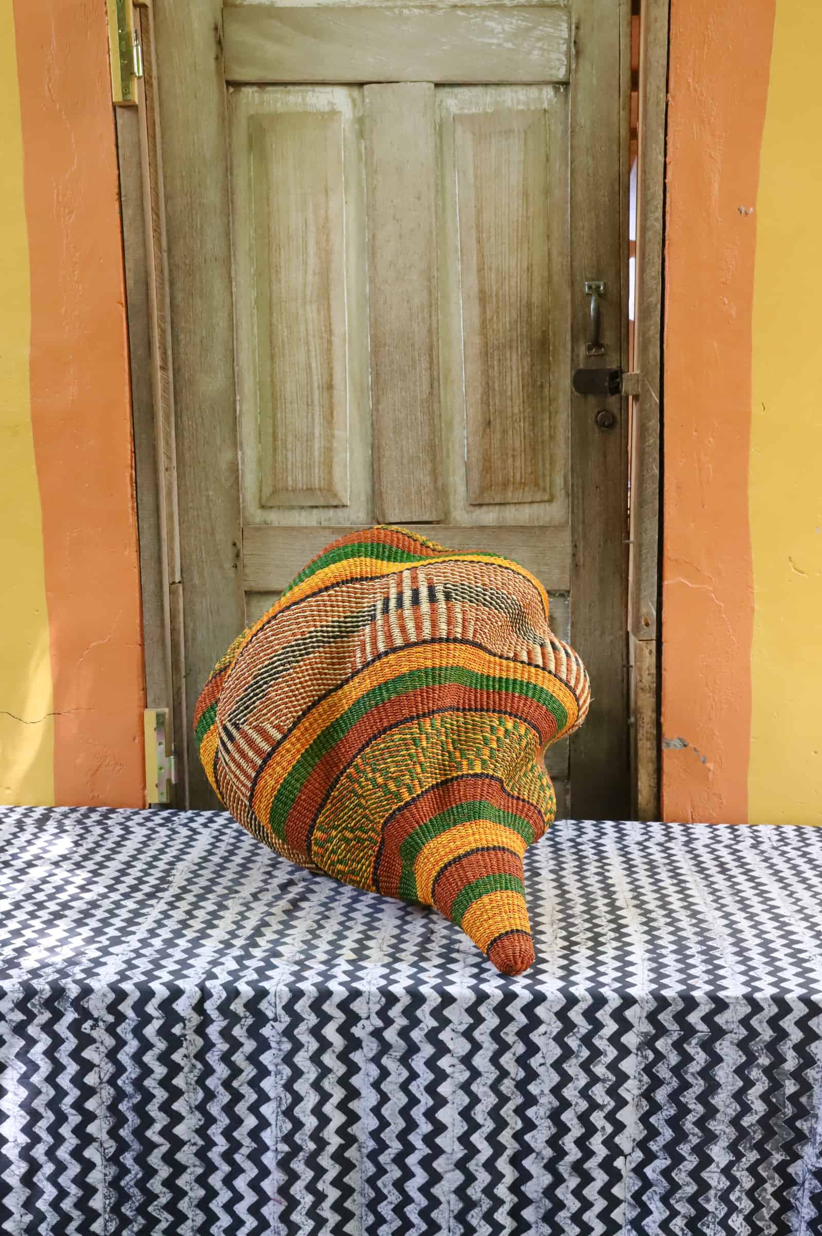 Handwoven Katayeli-tia art basket in amber and green, showing its conch-like spiral form, on a chevron cloth.