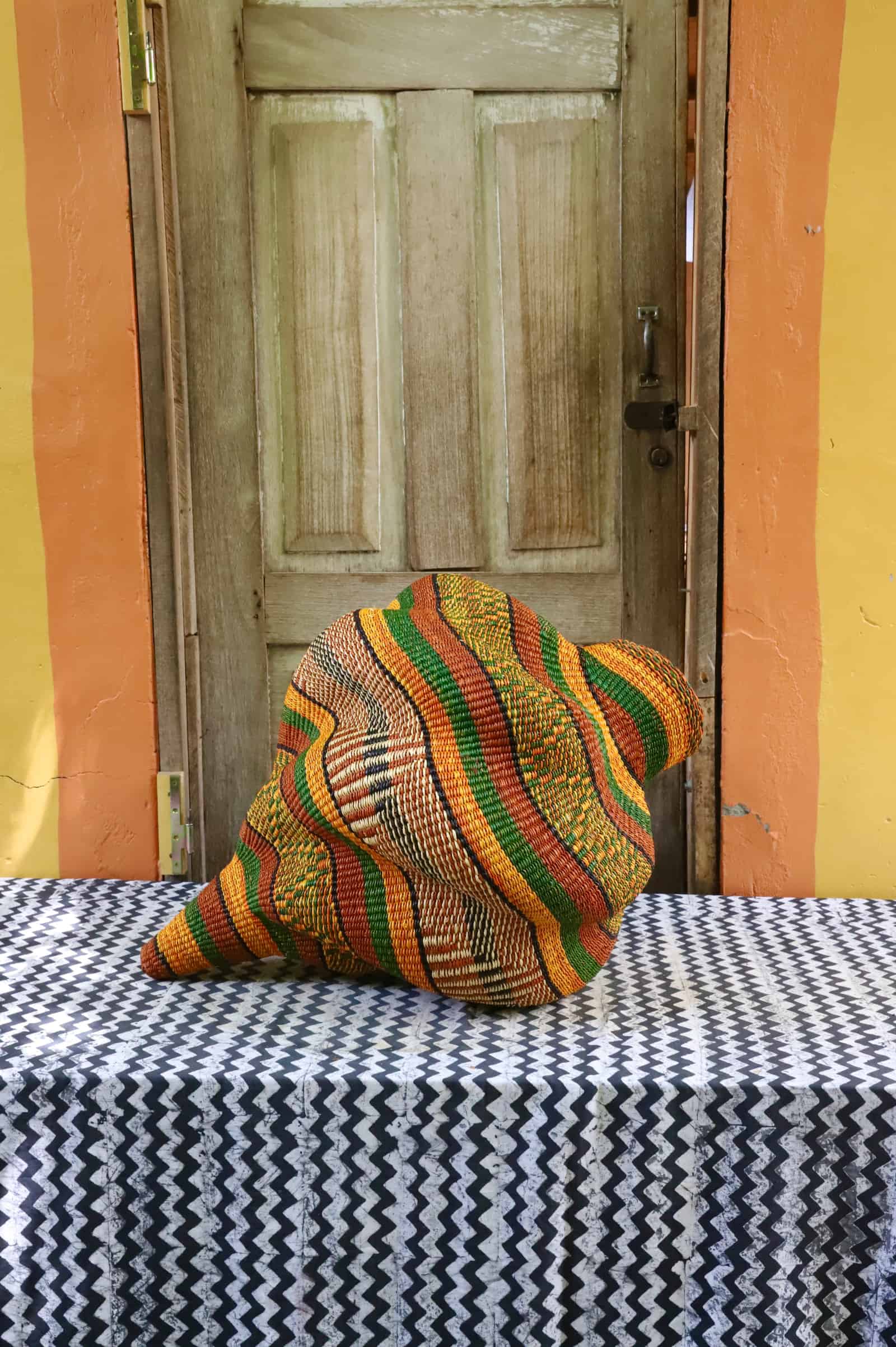 Handwoven Katayeli-tia art basket in amber and green stripes, its sculptural conch form on a black and white chevron cloth.