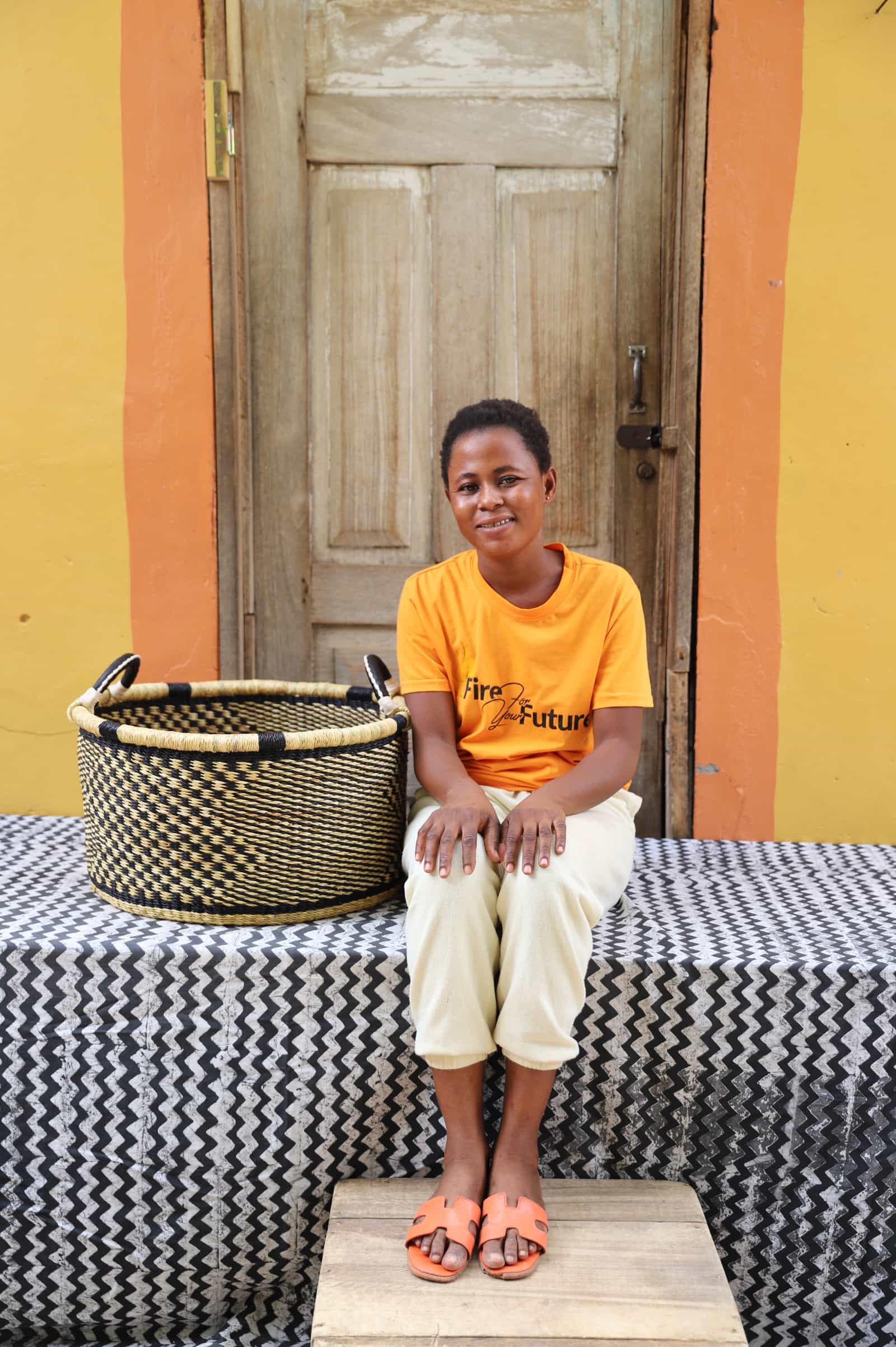 Woven Tub (Medium) with woman weaver handwoven in Ghana sitting on black and white cloth and wooden door in background

