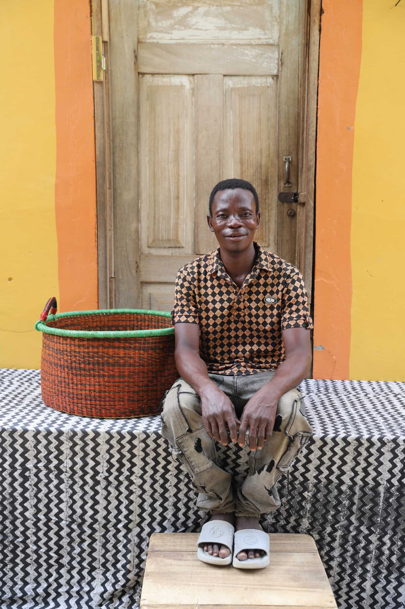 Woven Tub (Medium) with man weaver handwoven in Ghana sitting on black and white cloth and wooden door in background

