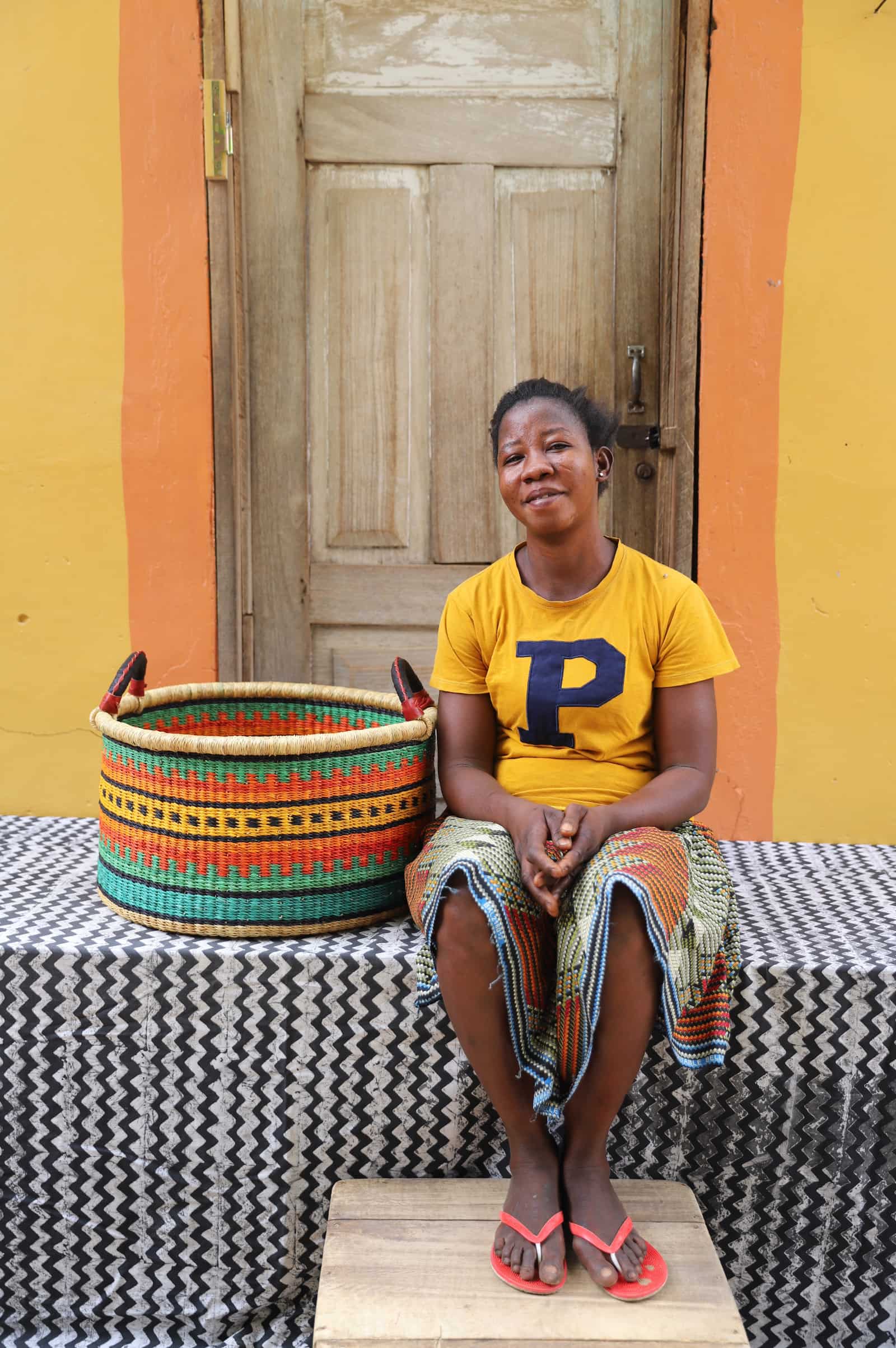 Woven Tub (Medium) with woman weaver handwoven in Ghana sitting on black and white cloth and wooden door in background

