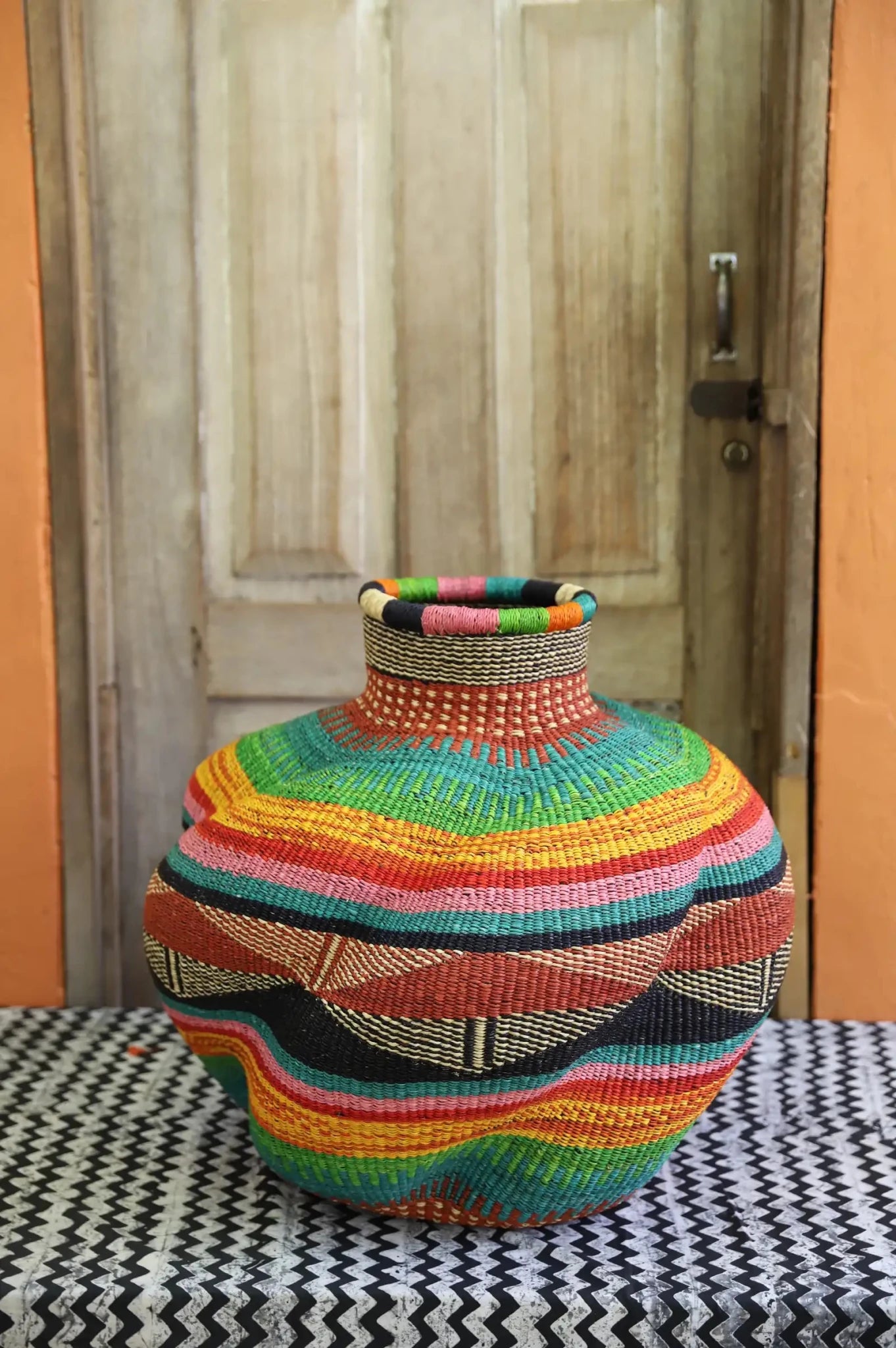 Colorful woven piece on a black and white patterned surface with a wooden door in the background.