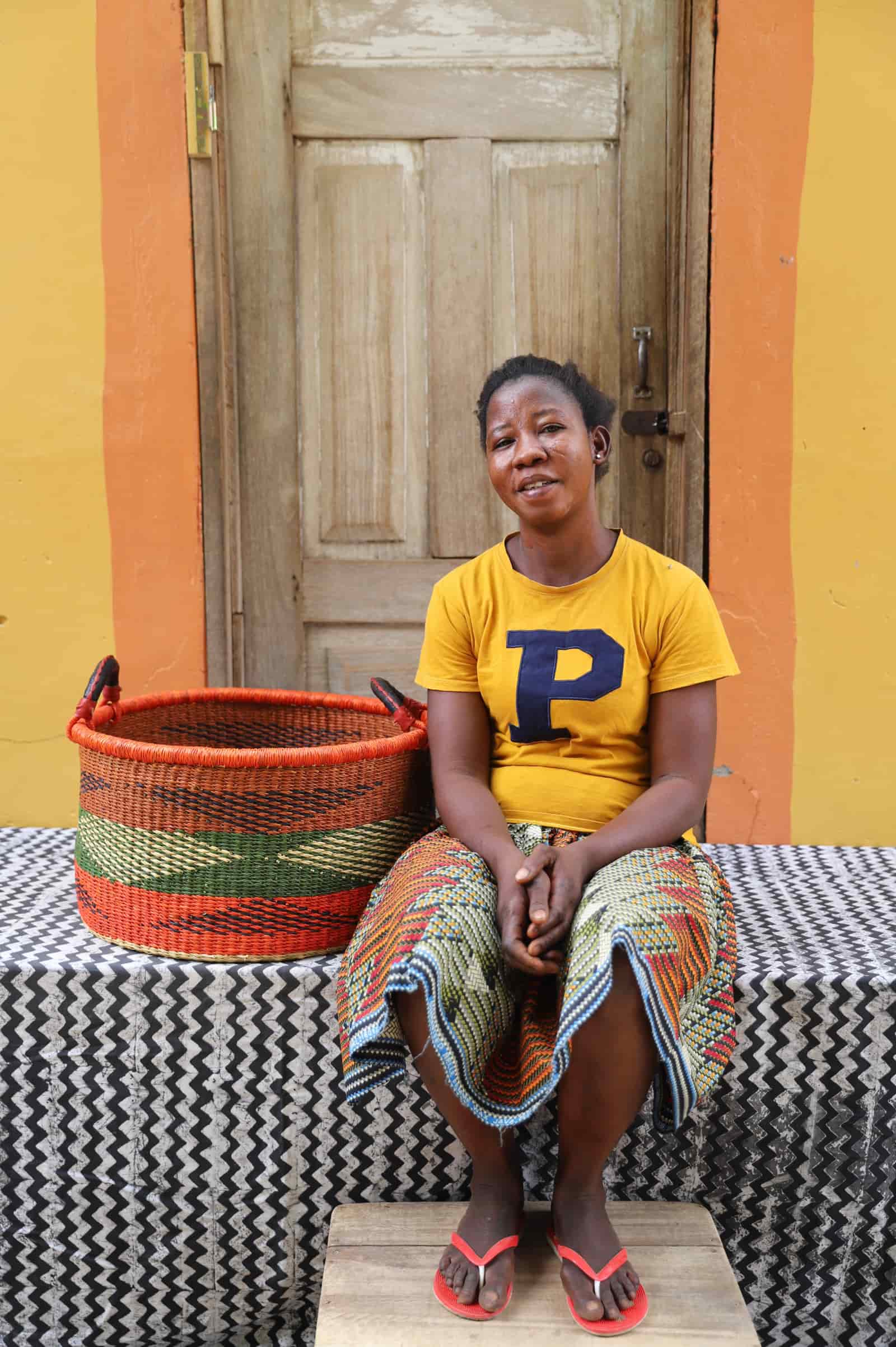 Woven Tub (Medium) with woman weaver handwoven in Ghana sitting on black and white cloth and wooden door in background

