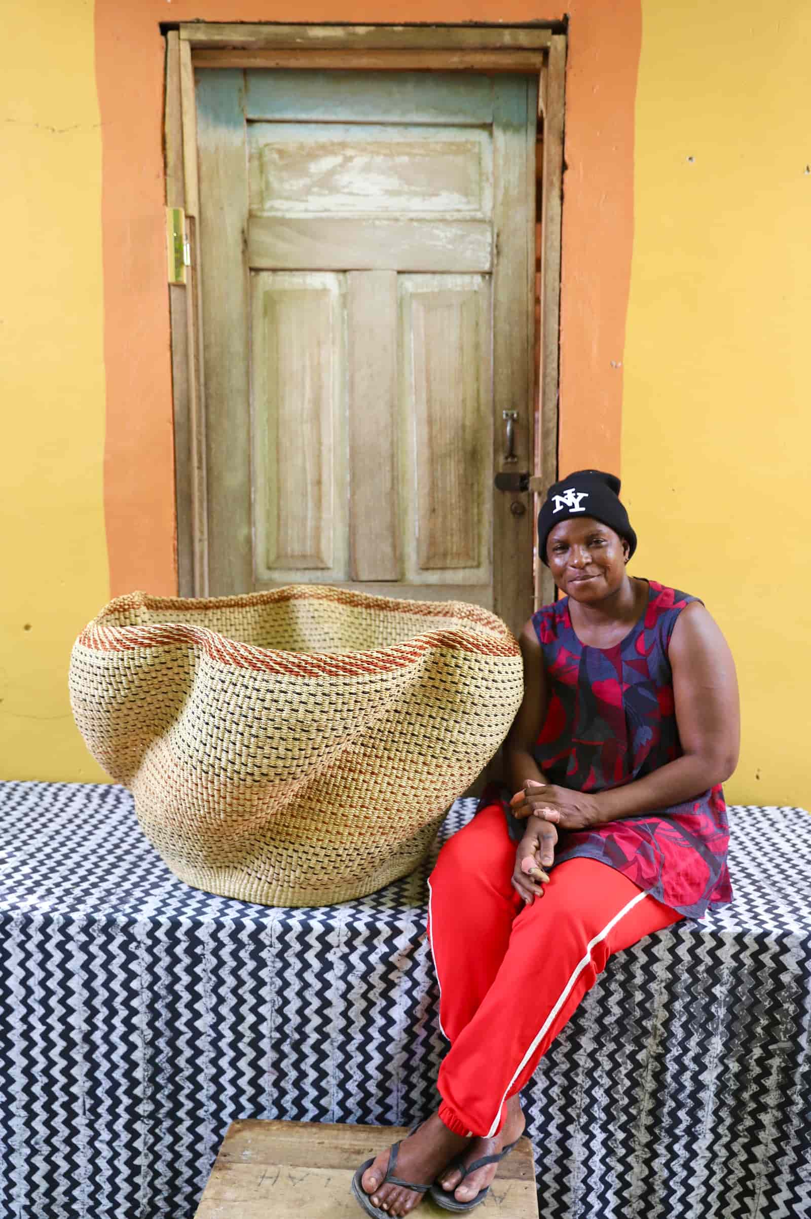 Woman seated beside a Giant Wave art basket in natural straw with a terracotta rim stripe, before a wooden door.