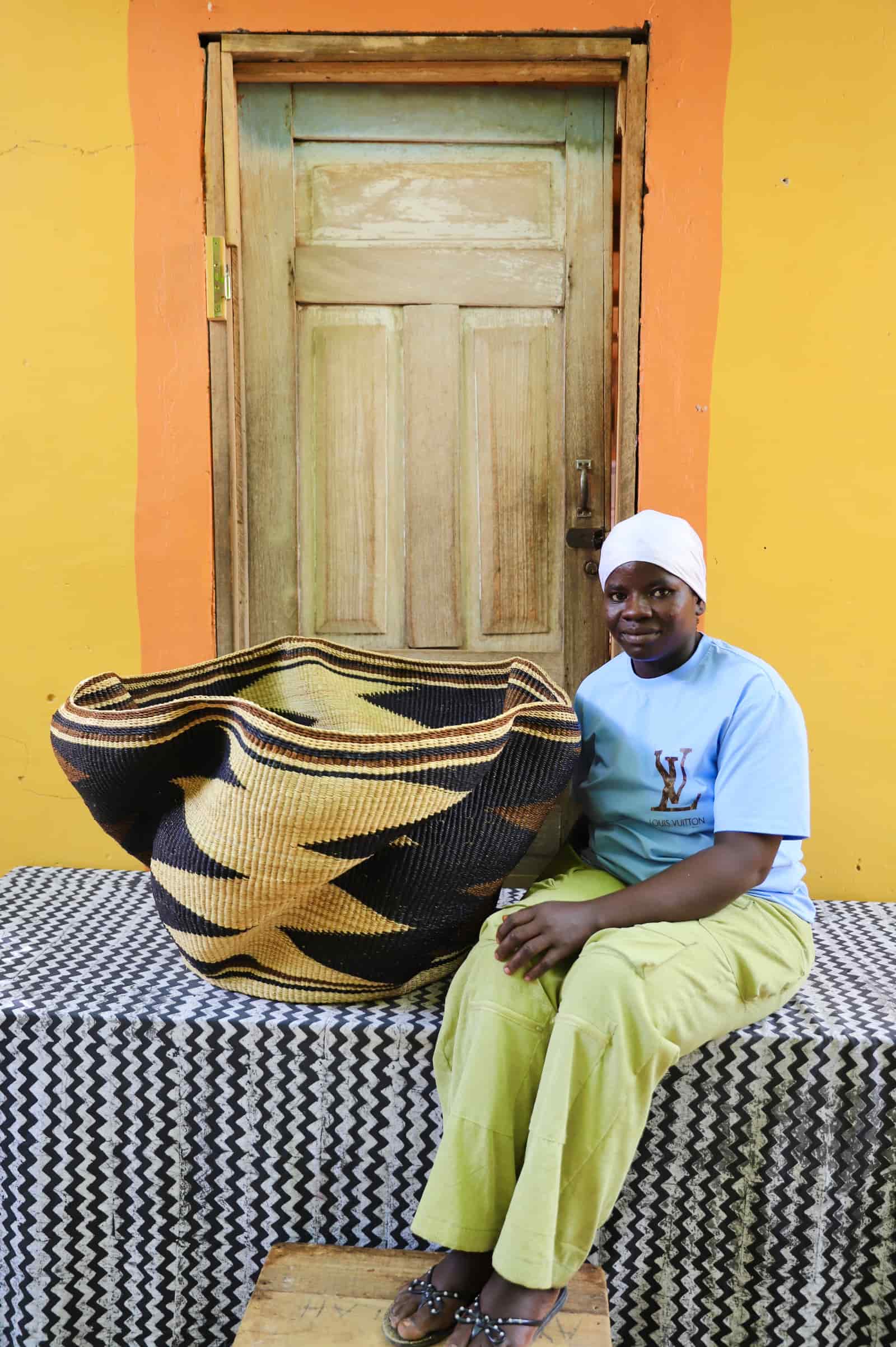 Woman seated beside a Giant Wave art basket in natural straw with bold black wave bands, before a wooden door.