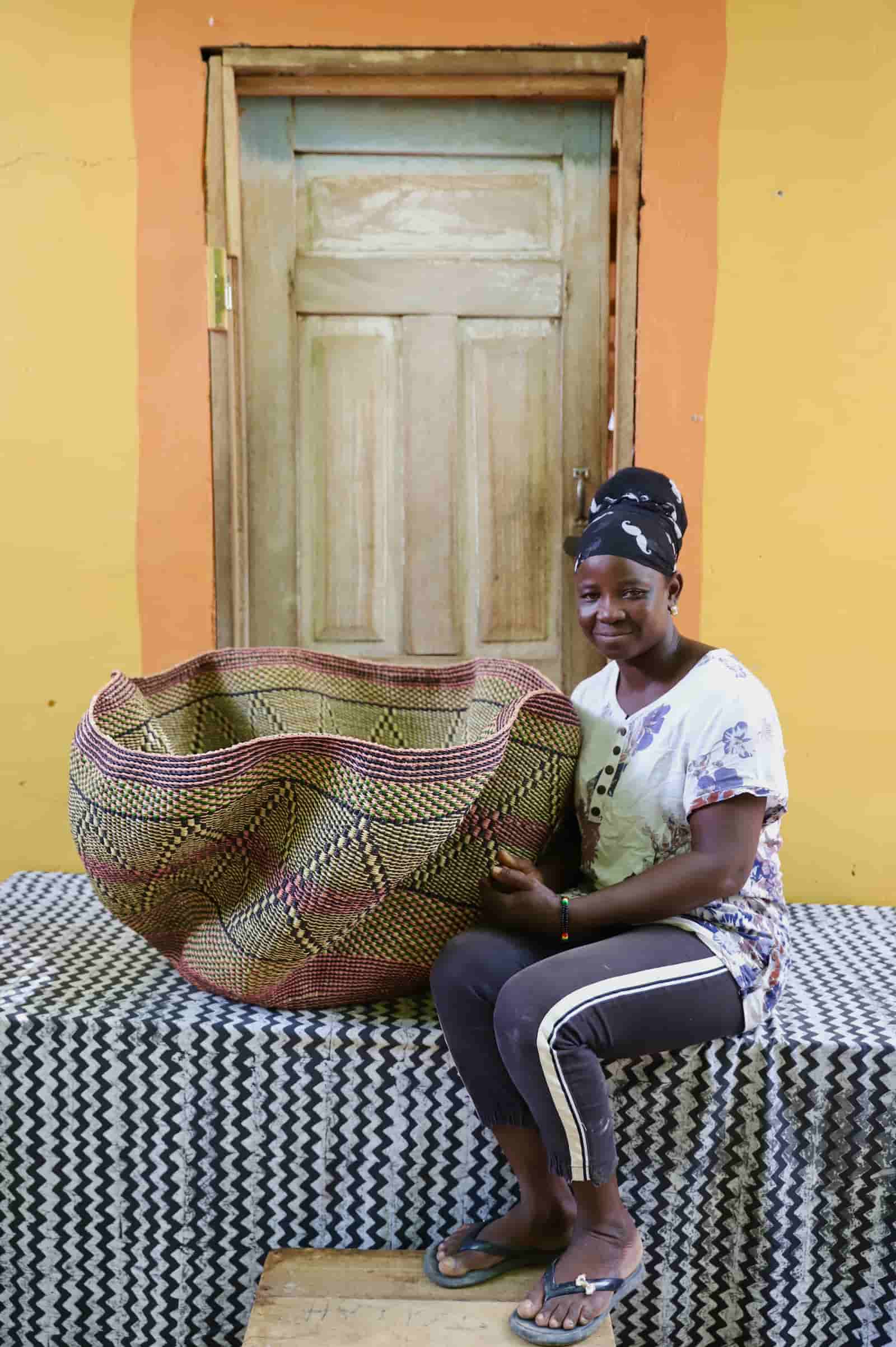 Woman seated beside a Giant Wave art basket with an olive green and dark brown diamond pattern, before a wooden door.