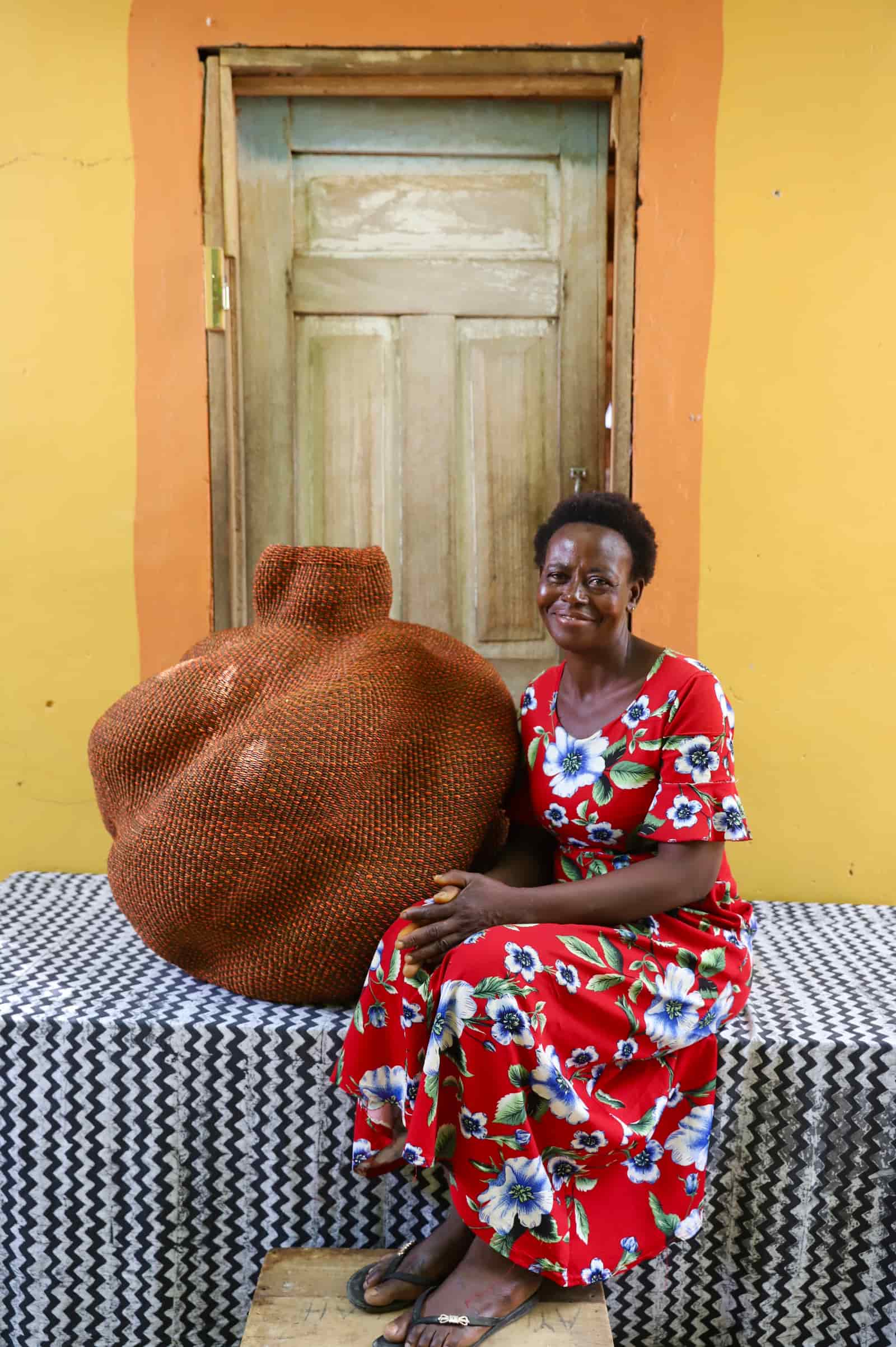 Woman in a red floral dress seated beside a large rounded terracotta woven basket, against a wooden door