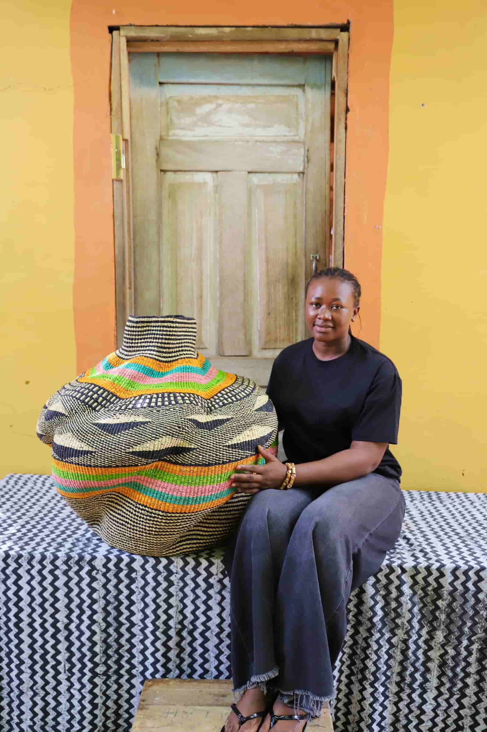 Woman seated beside a rounded woven basket with bold orange, teal and pink bands on black pattern, against a wooden door