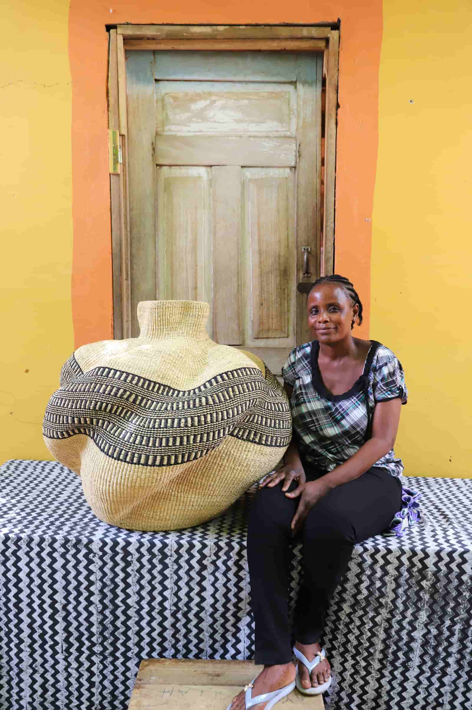 Woman seated beside a bulbous woven basket in natural straw with black and cream geometric band, in front of a wooden door