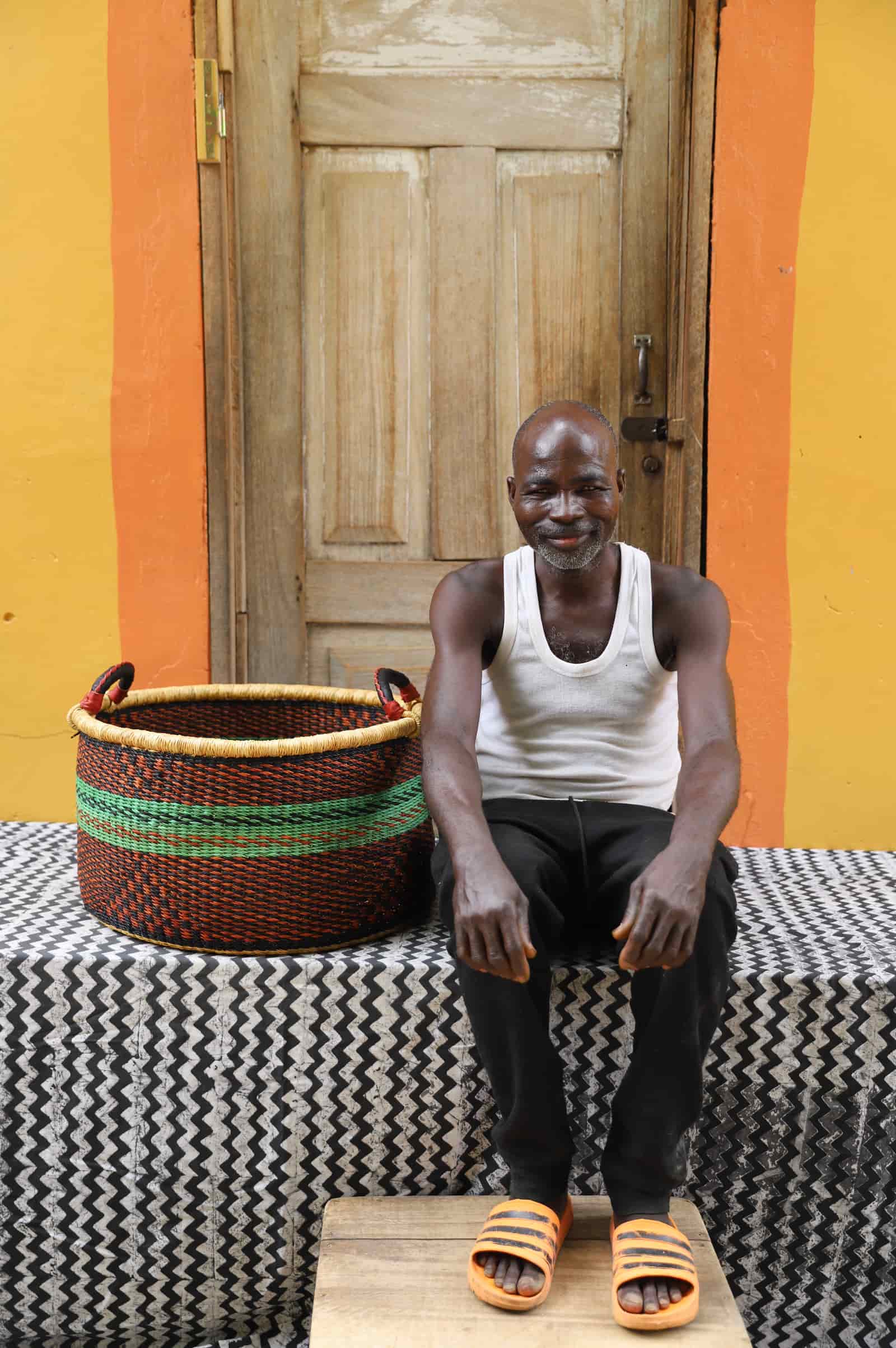 Woven Tub (Medium) with man weaver handwoven in Ghana sitting on black and white cloth and wooden door in background