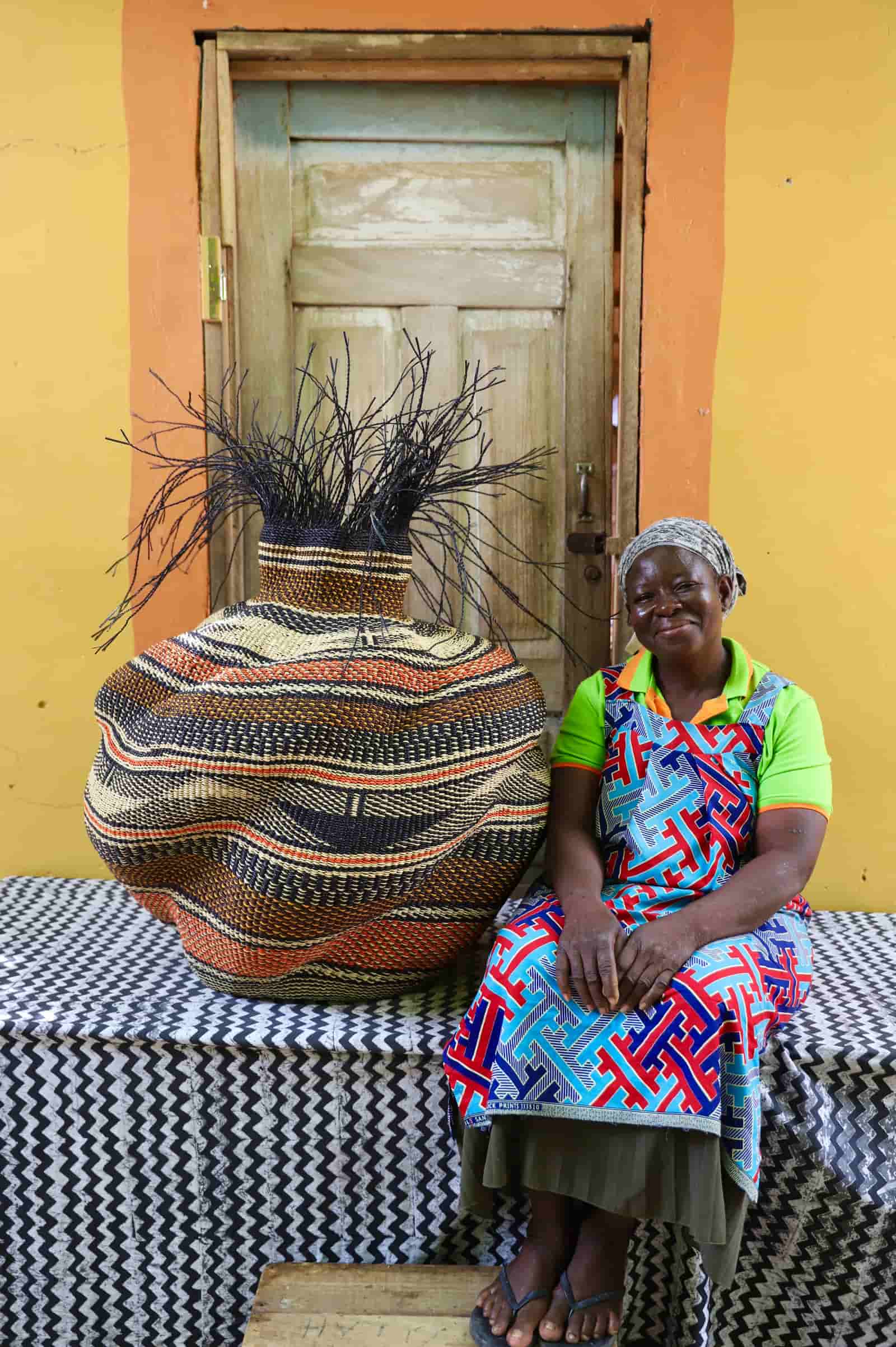 Woman in a colourful patterned apron sitting beside the large Tua-Tia baobab art basket in dark and terracotta stripes.