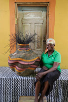 Woman sitting next to a large woven basket in front of a yellow wall with a wooden door.