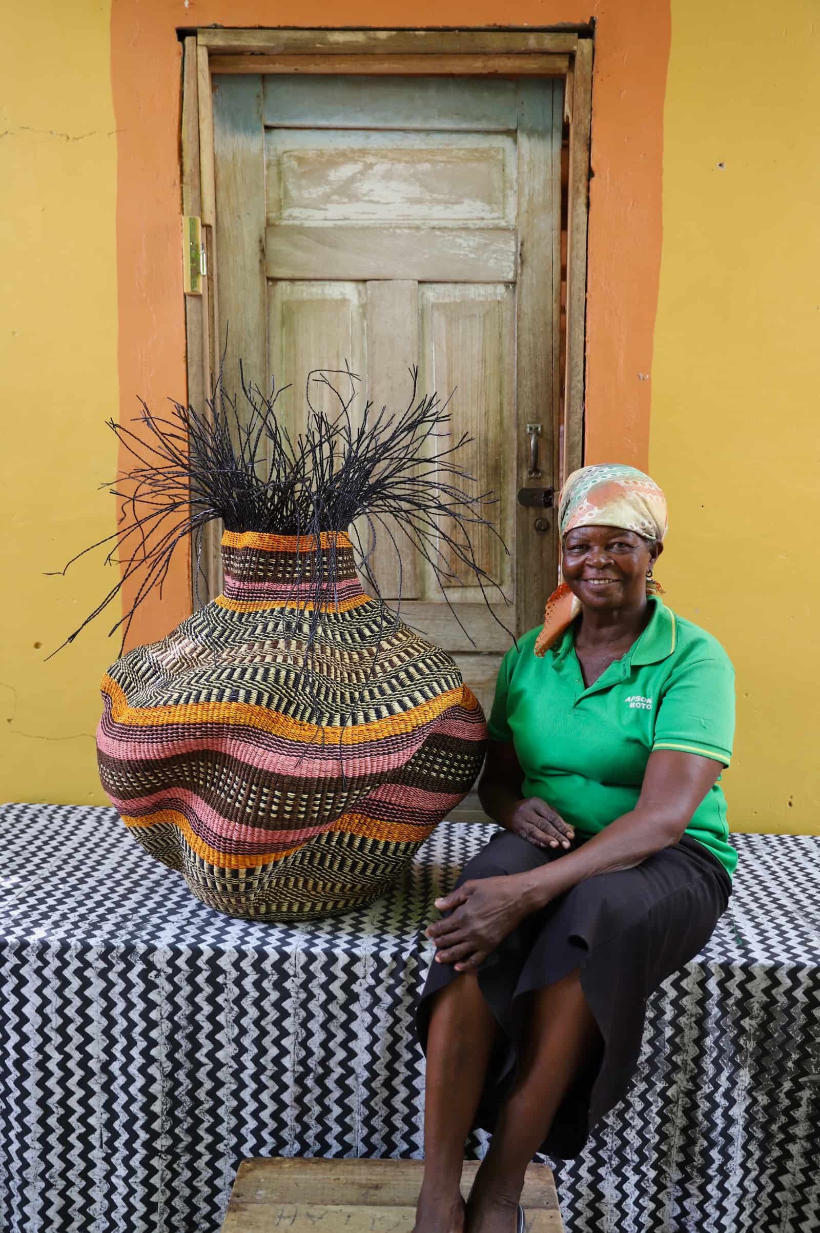 Woman sitting next to a large woven basket in front of a yellow wall with a wooden door.