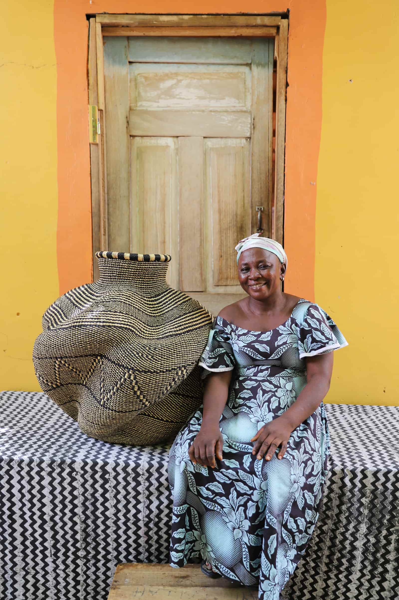 Woman seated beside a large handwoven De-engo art basket in natural straw and black geometric pattern, before a wooden door.