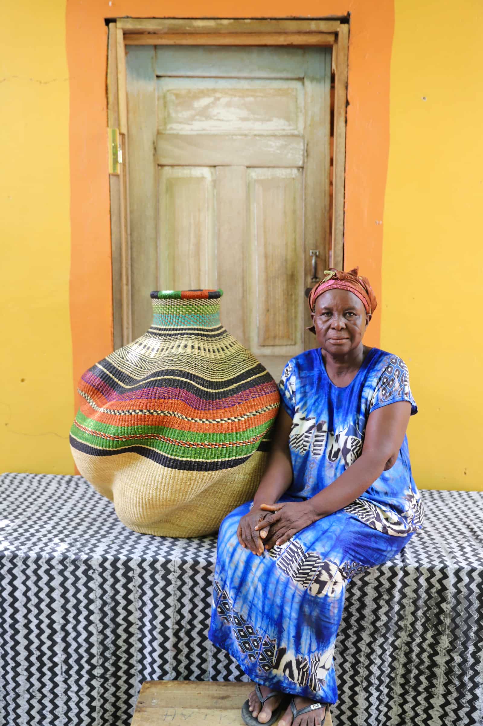Woman seated beside a large handwoven De-engo art basket in terracotta, green and black stripes, before a wooden door.