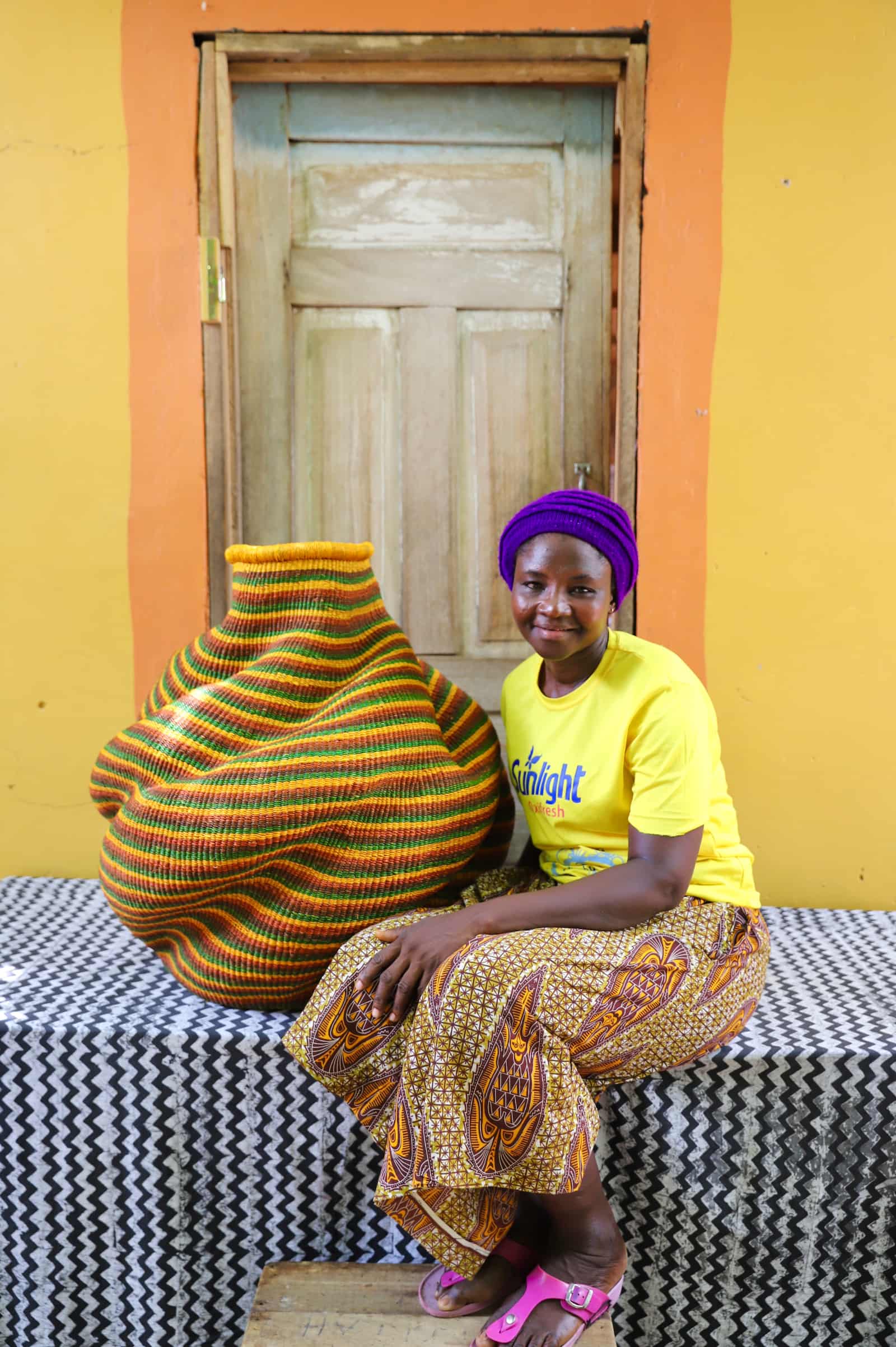Woman seated beside a large handwoven De-engo art basket in amber, green and terracotta stripes, before a wooden door.