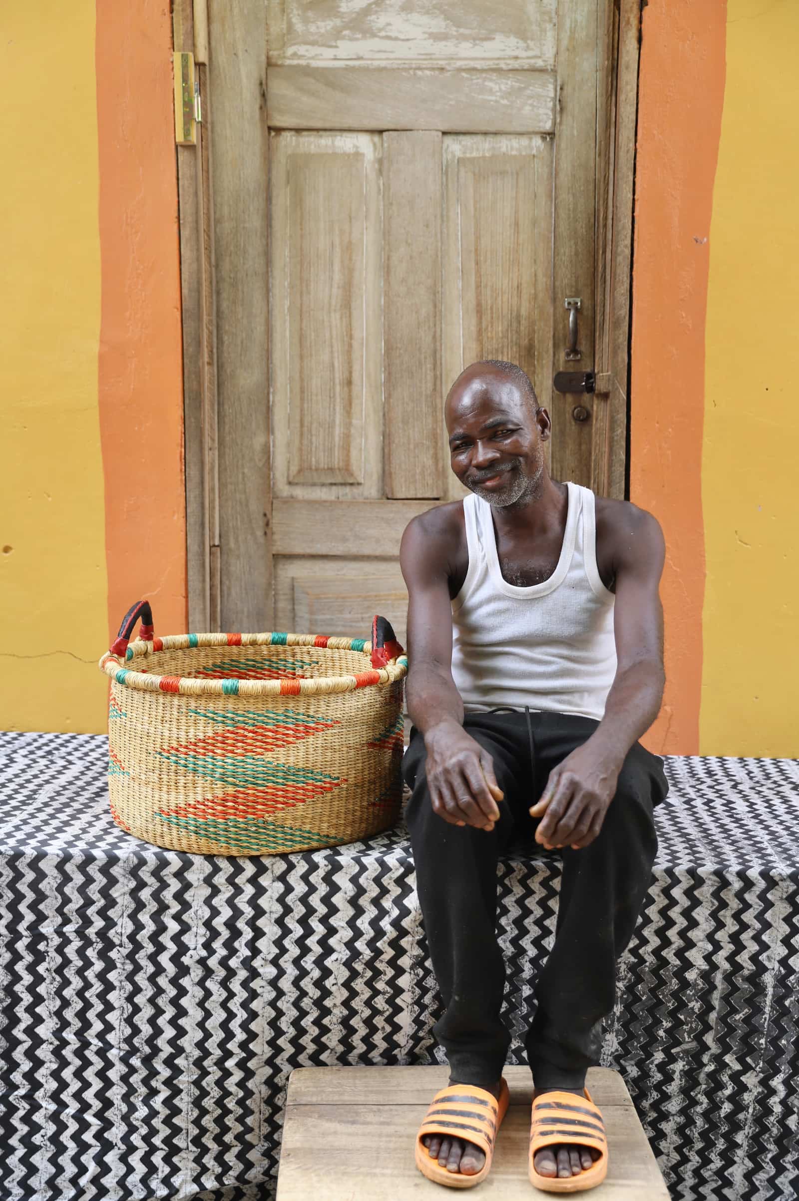 Woven Tub (Small) with man weaver handwoven in Ghana sitting on black and white cloth and wooden door in background