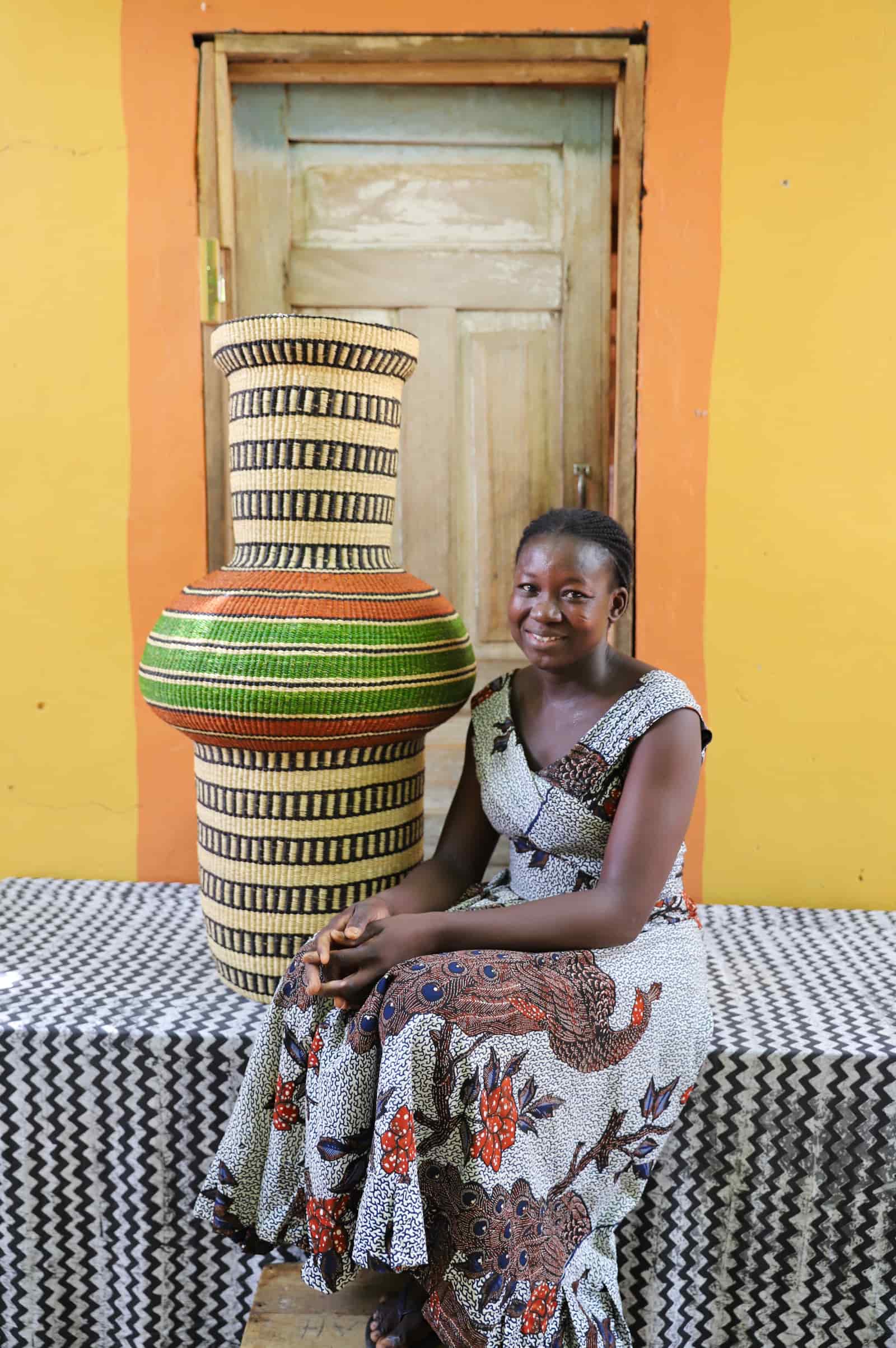 Woman seated beside a tall Dugla art basket in natural straw with black stripes and a green band, before a wooden door.