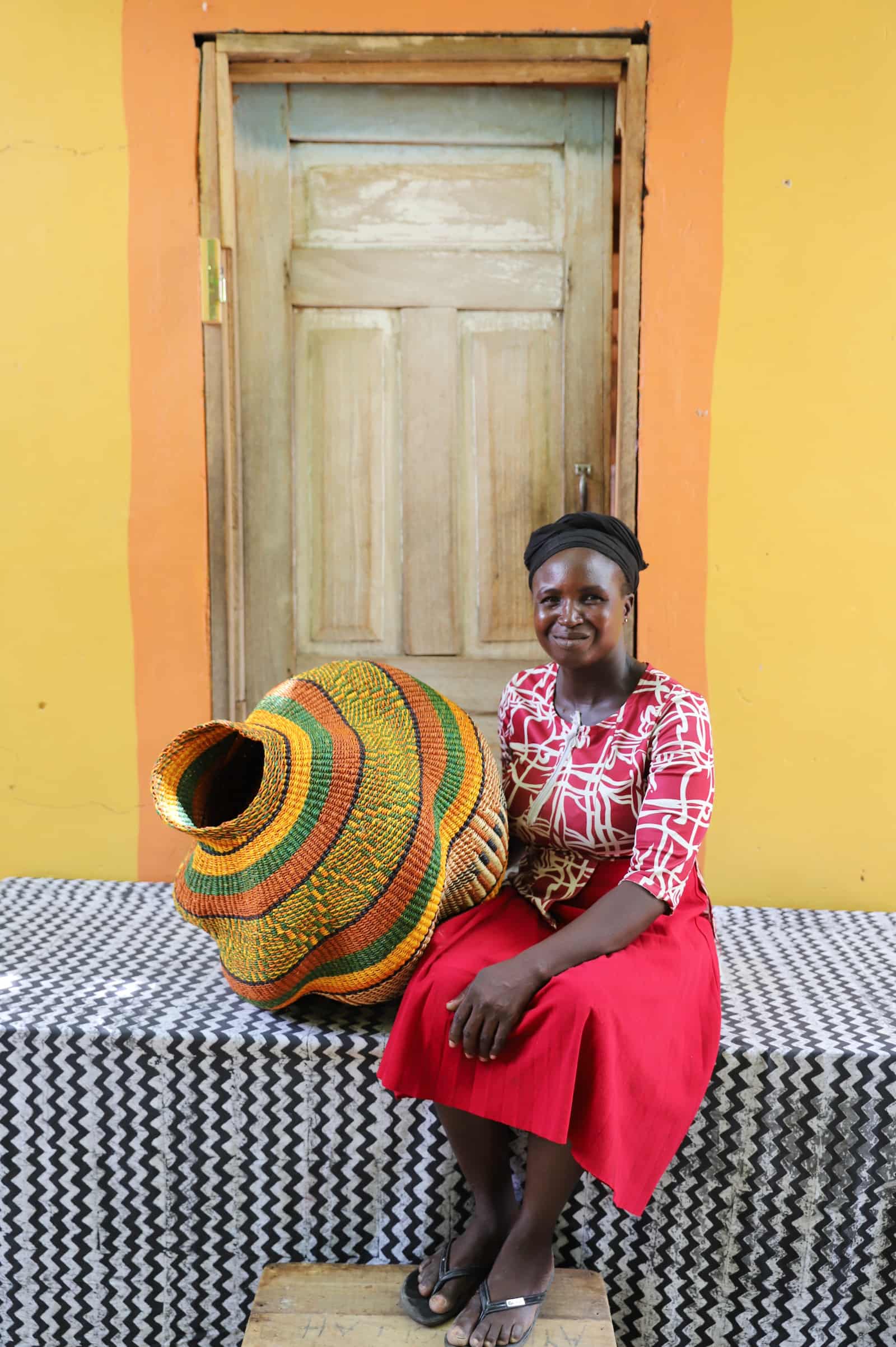Woman seated beside a handwoven Katayeli-tia art basket in amber and green stripes, before a wooden door with a yellow wall.