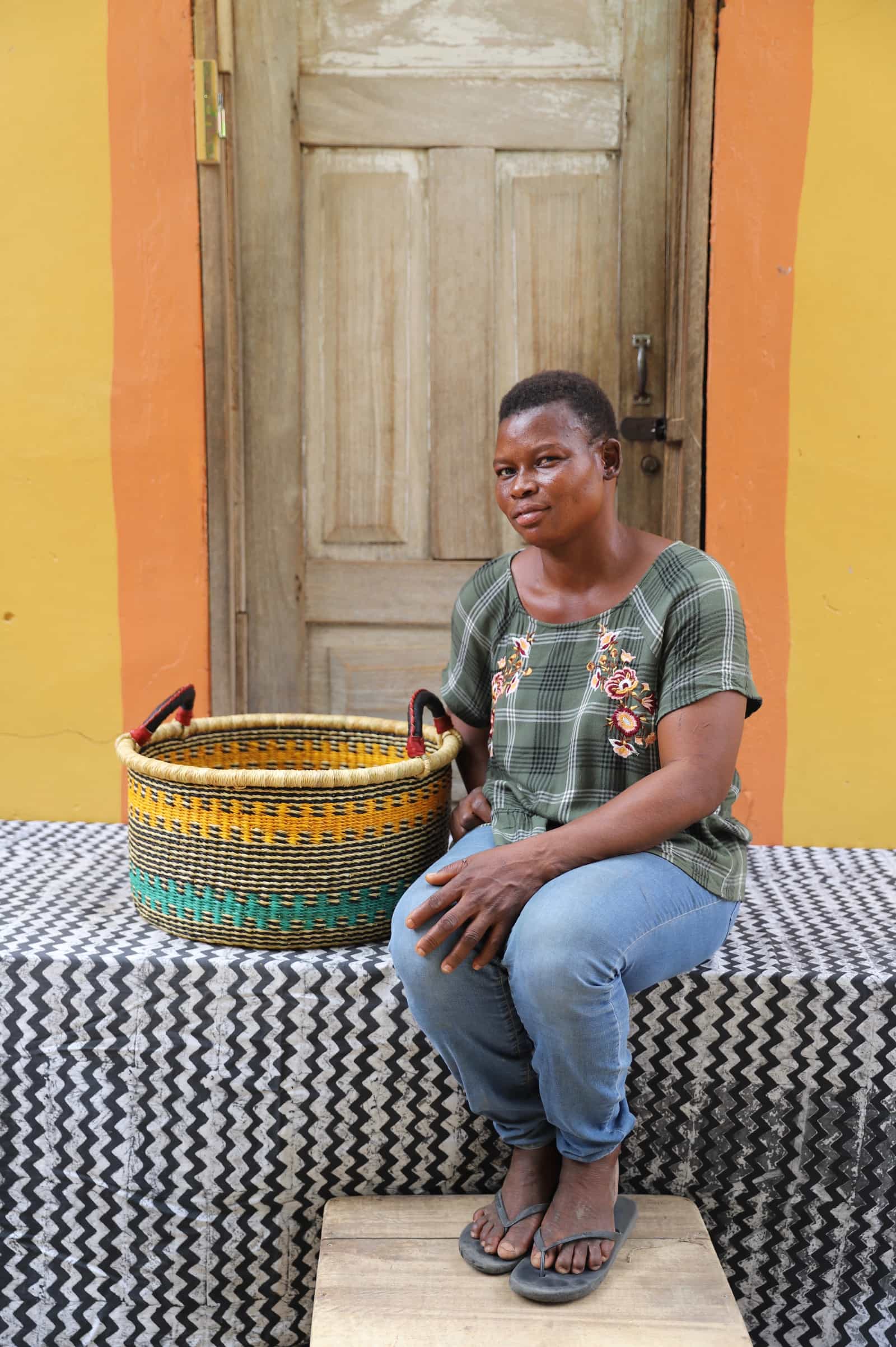 Woven Tub (Small) with woman weaver handwoven in Ghana sitting on black and white cloth and wooden door in background


