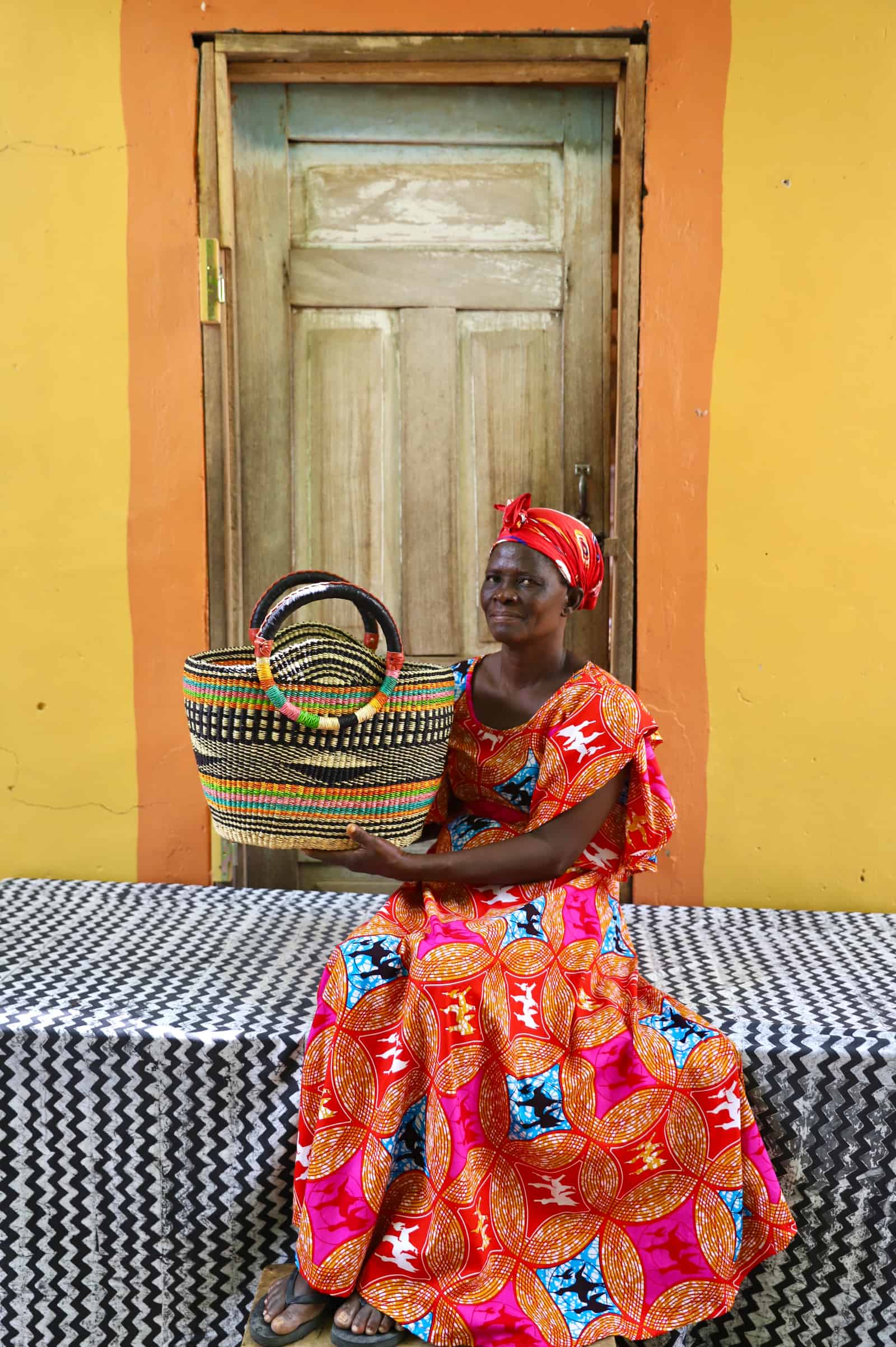 Woman in colorful traditional attire holding a woven basket against a yellow wall.