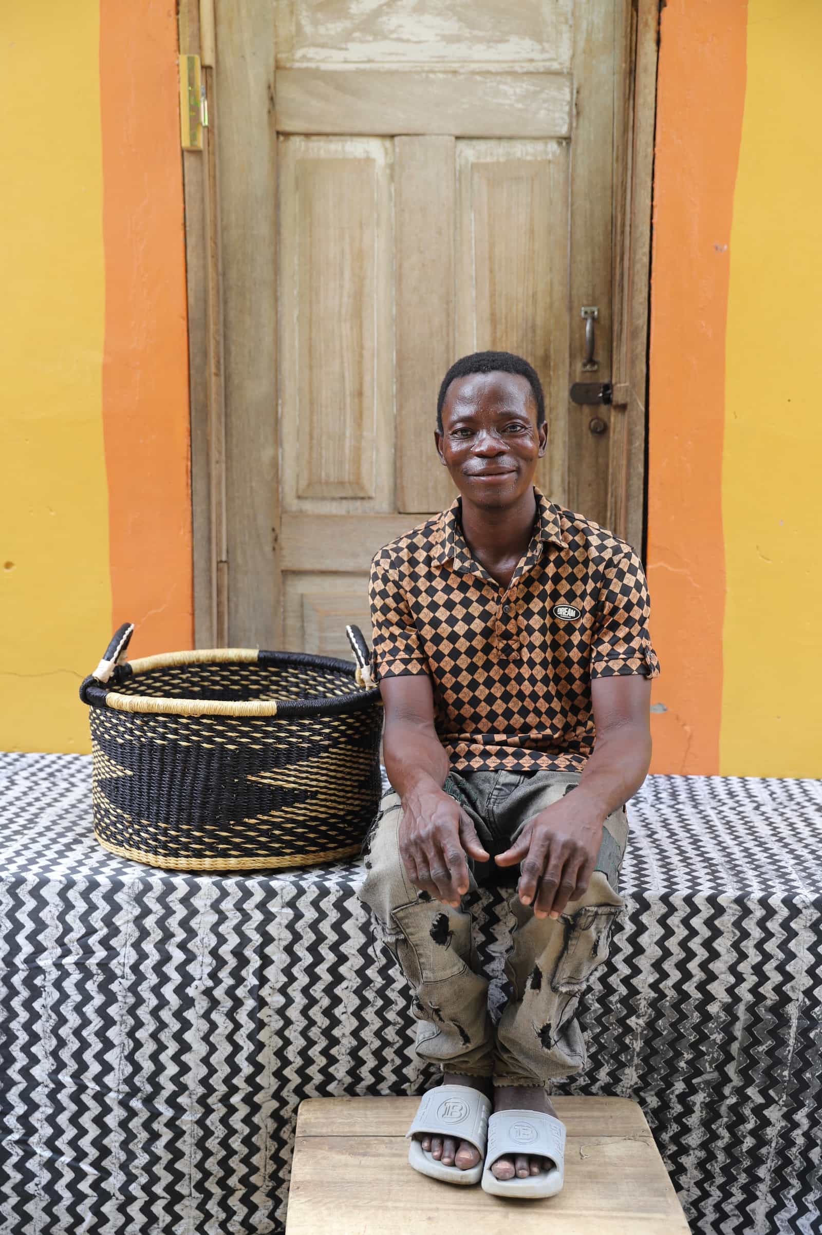 Woven Tub (Small) with man weaver handwoven in Ghana sitting on black and white cloth and wooden door in background

