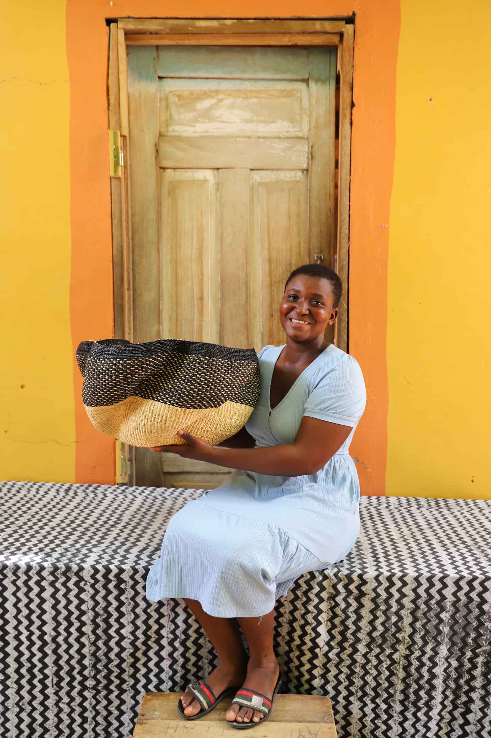 Woman holding a woven basket in front of a yellow wall with a wooden door.