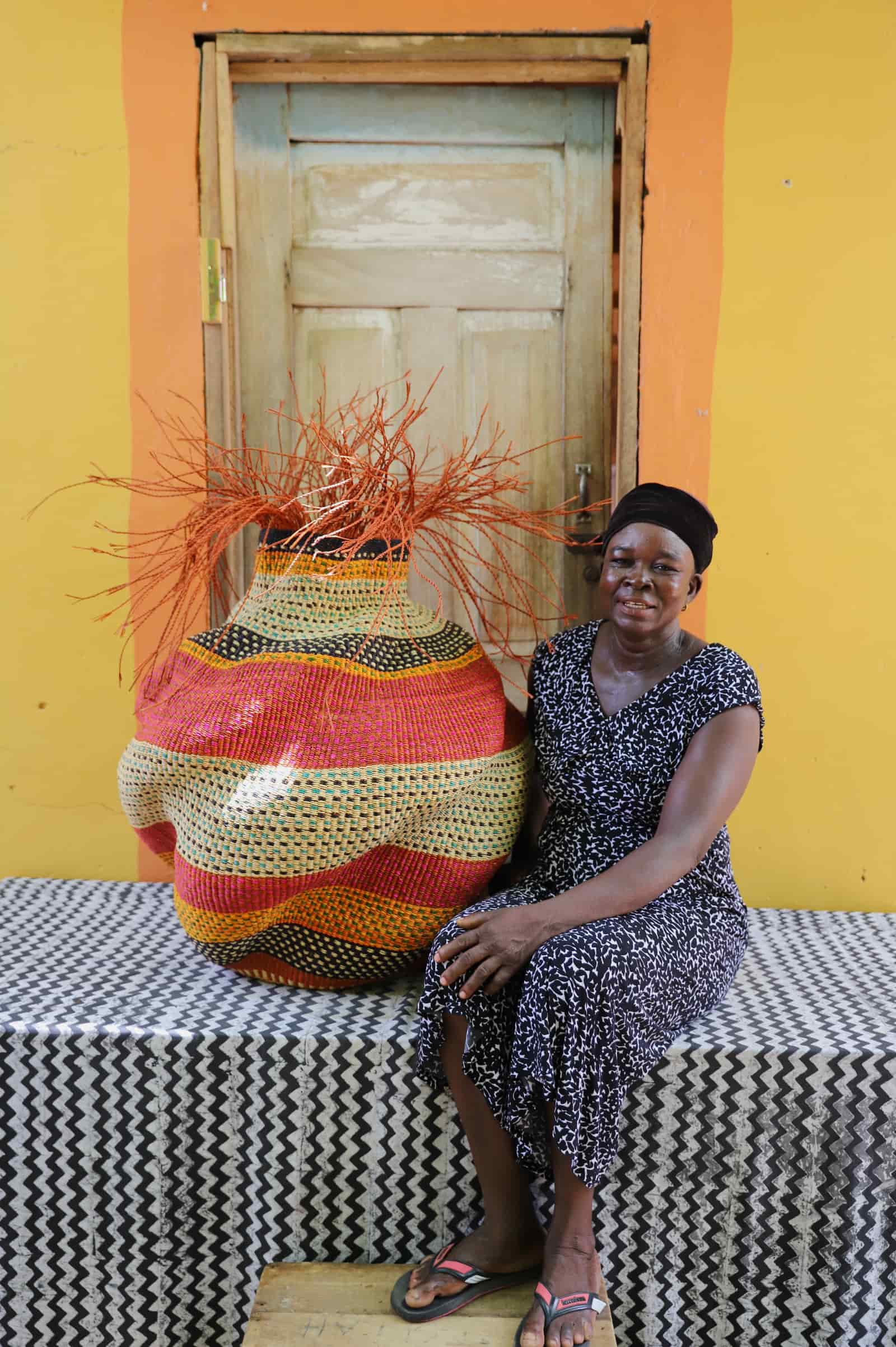 Woman sitting beside the large Tua-Tia baobab art basket in red and natural stripes against an orange wall.