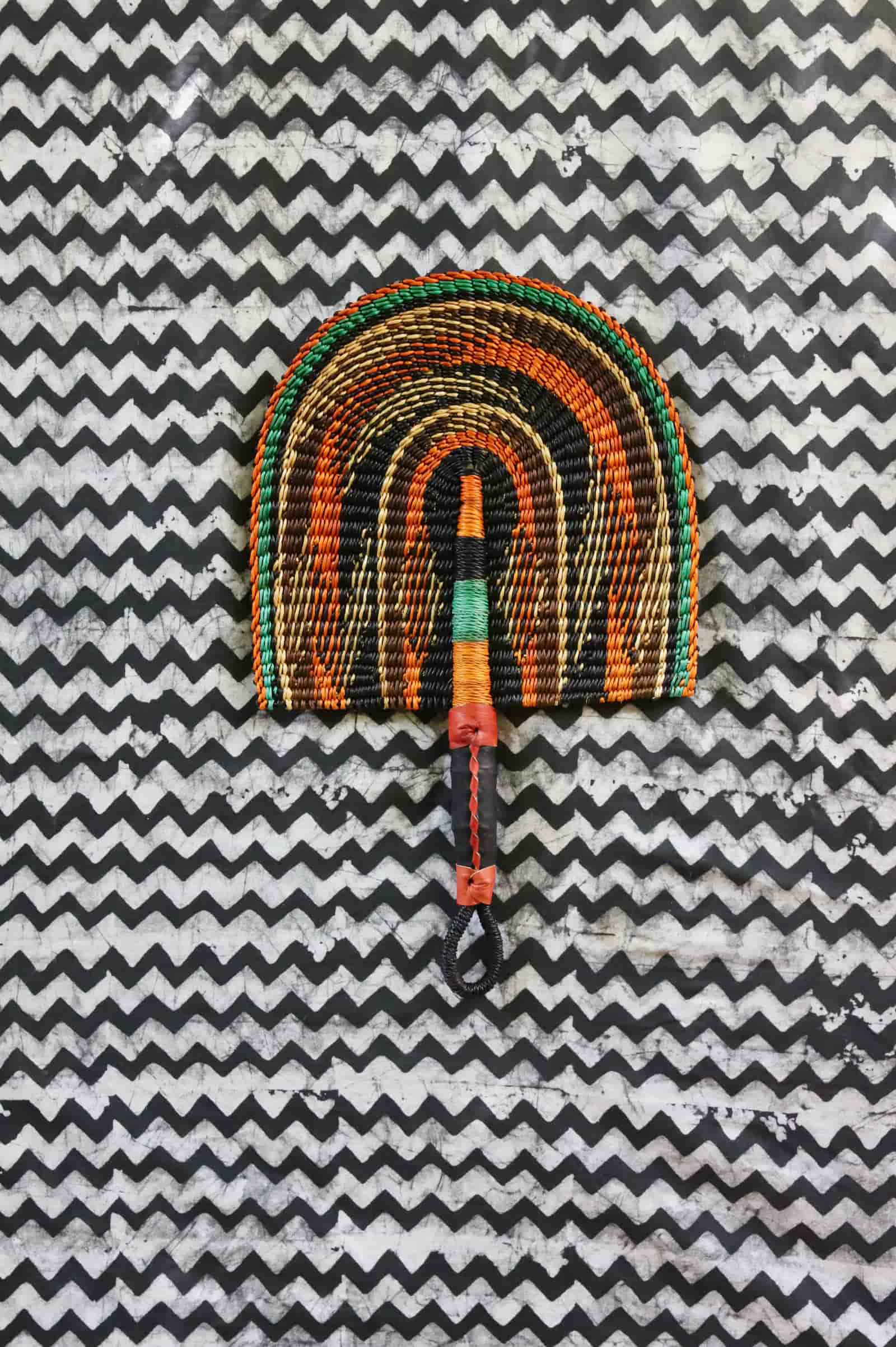 Handwoven Bolga fan in dark brown with amber and teal arch outlines, woven handle, on a black and white chevron cloth.