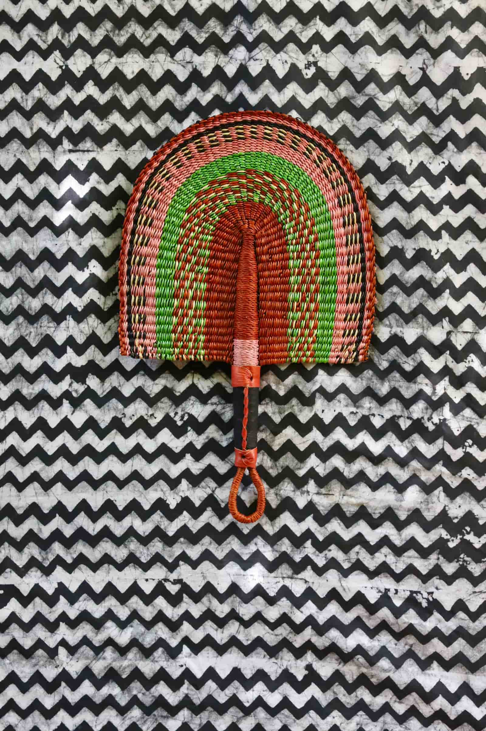 Handwoven Bolga fan in terracotta, green and pink concentric arch pattern, tan leather handle, on a chevron cloth.