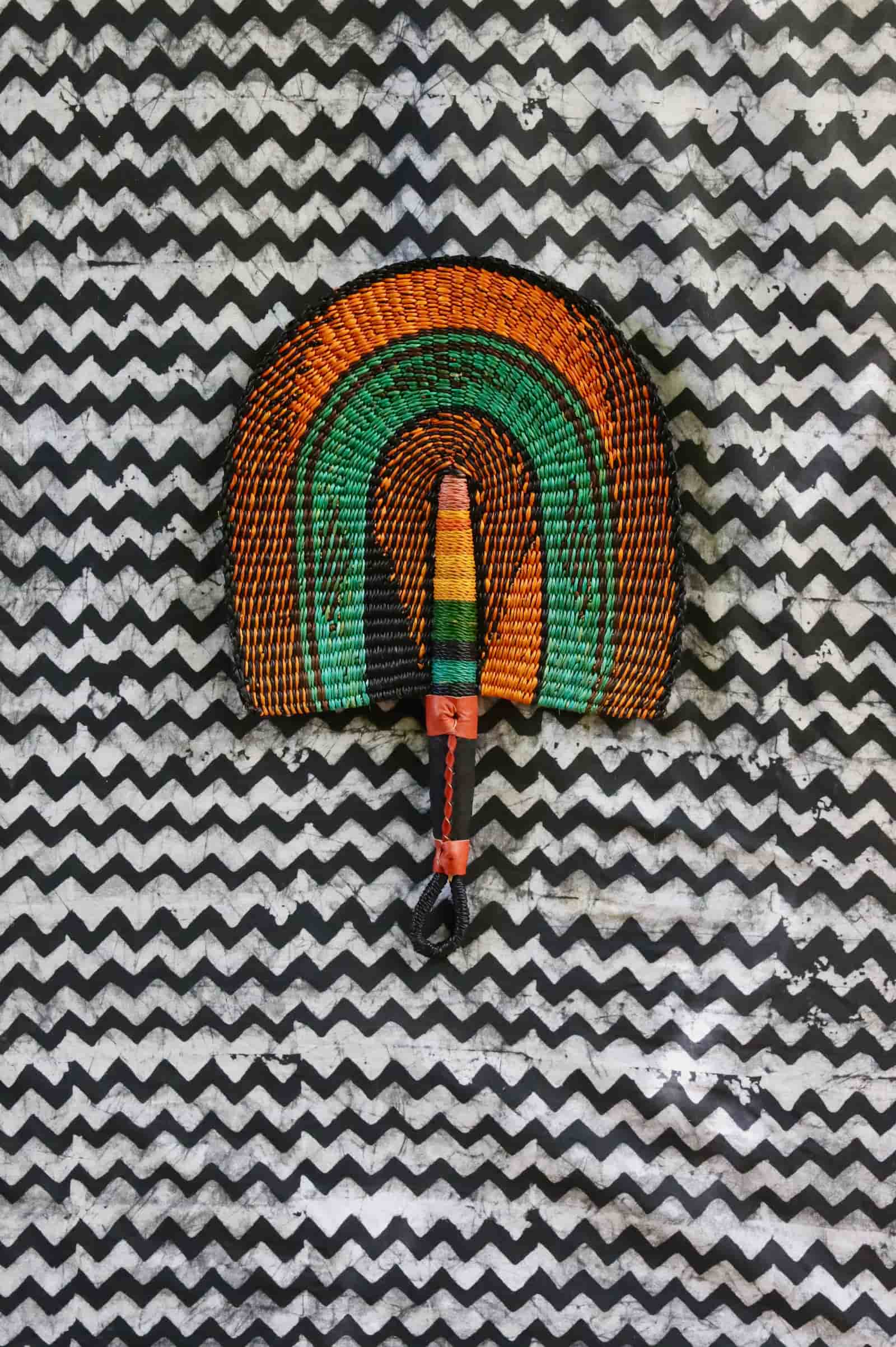 Handwoven Bolga fan with amber outer arches and teal inner arch, dark leather handle, on a black and white chevron cloth.