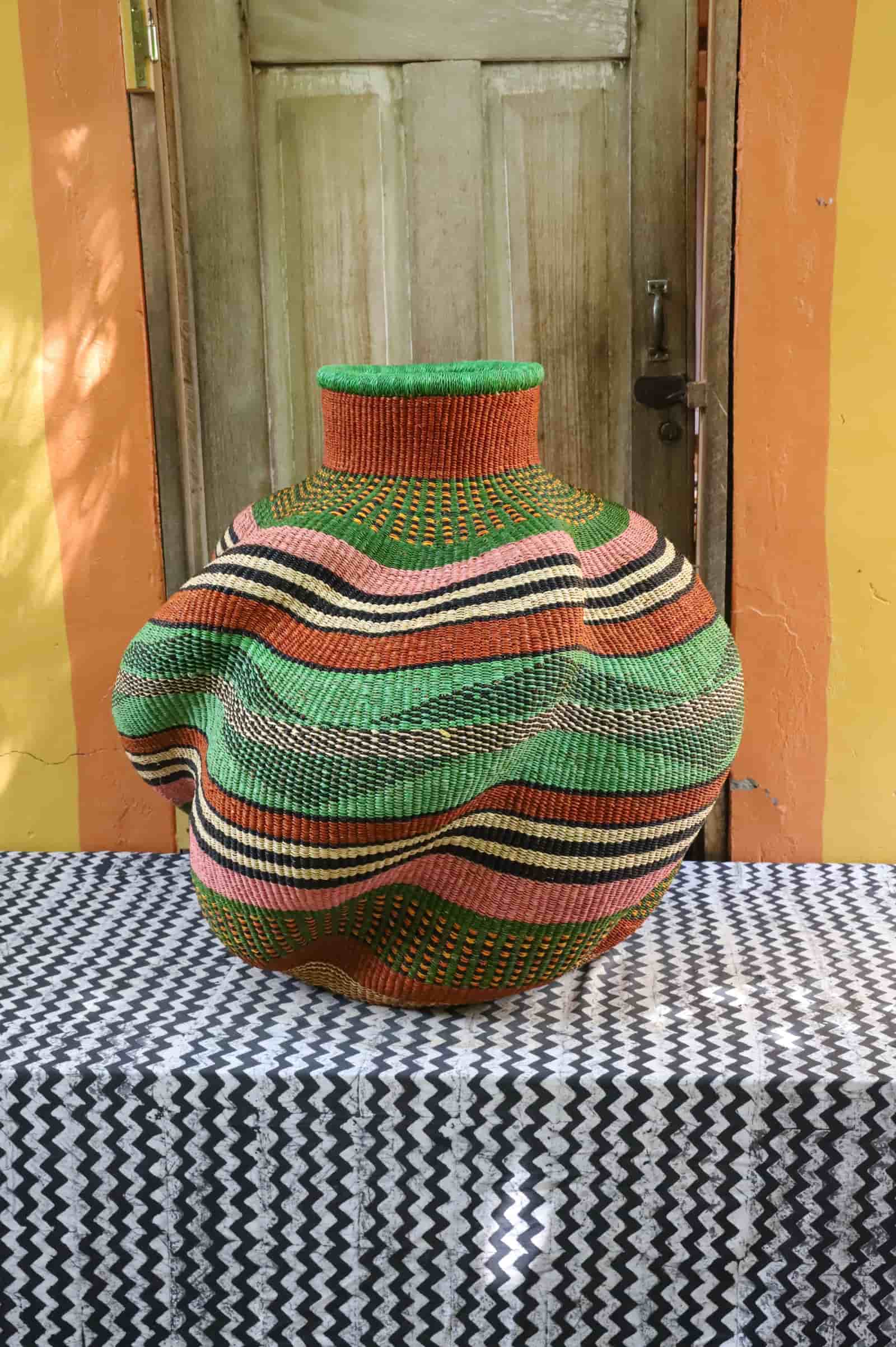 Woman holding a small handwoven De-engo art basket in natural straw and dark red stripes, before a wooden door.