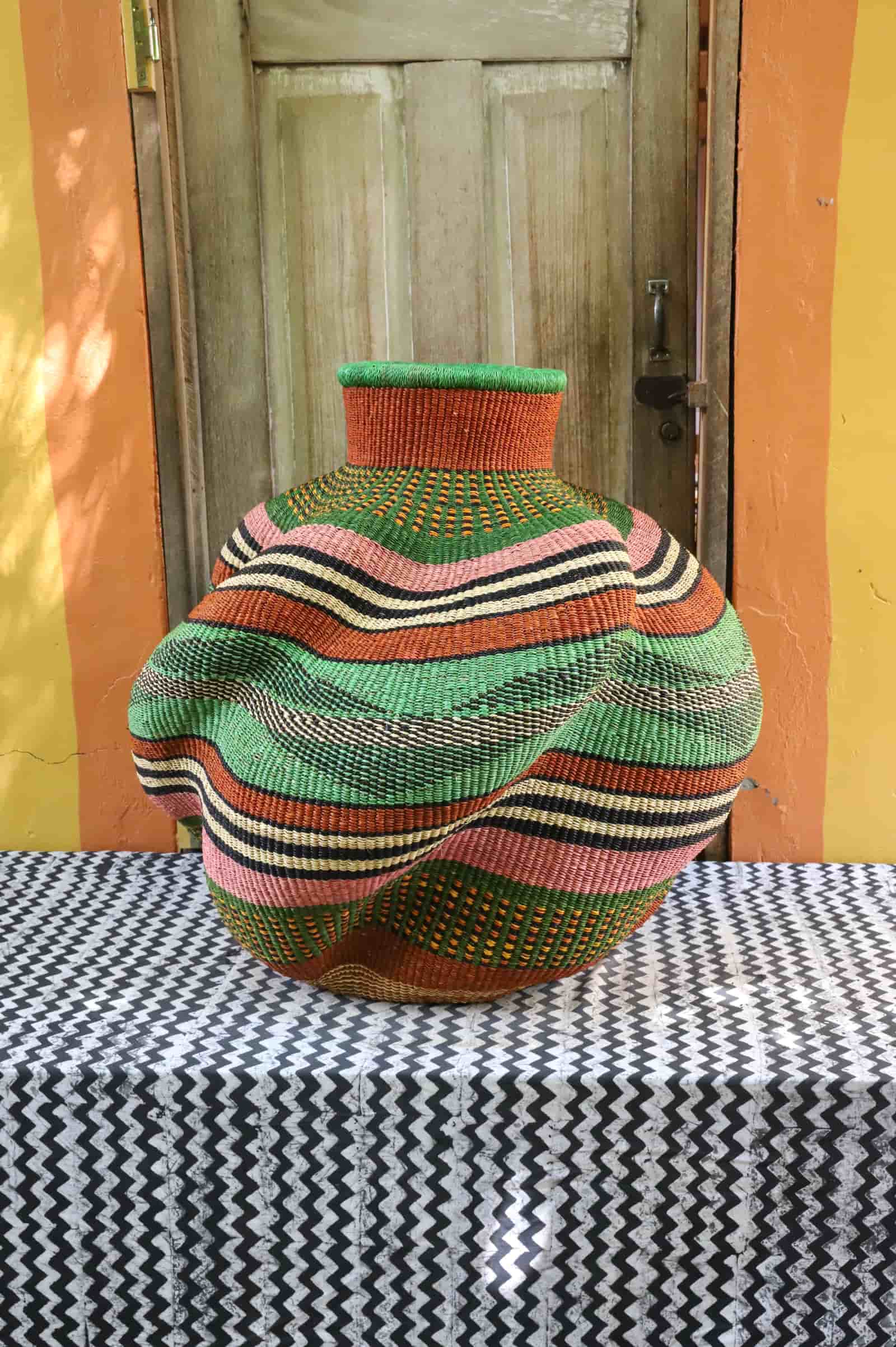Side view of a large handwoven De-engo art basket in green, terracotta and pink stripes, on a chevron cloth.