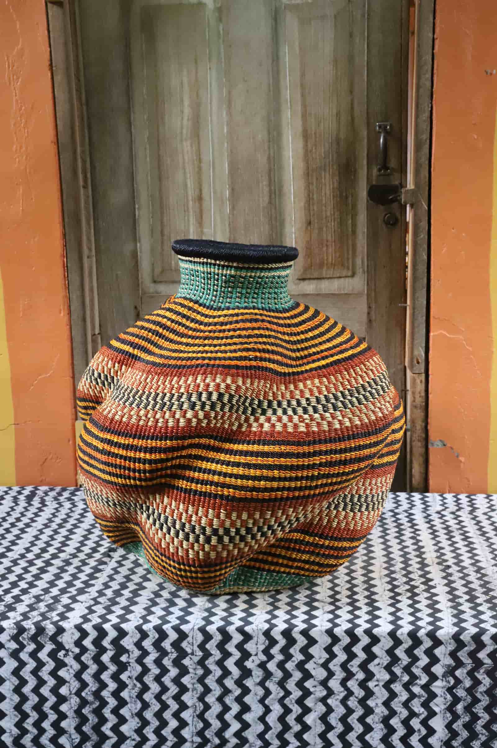 Handwoven De-engo art basket with a lobed gourd form and stripes in amber, terracotta and teal, on a chevron cloth.