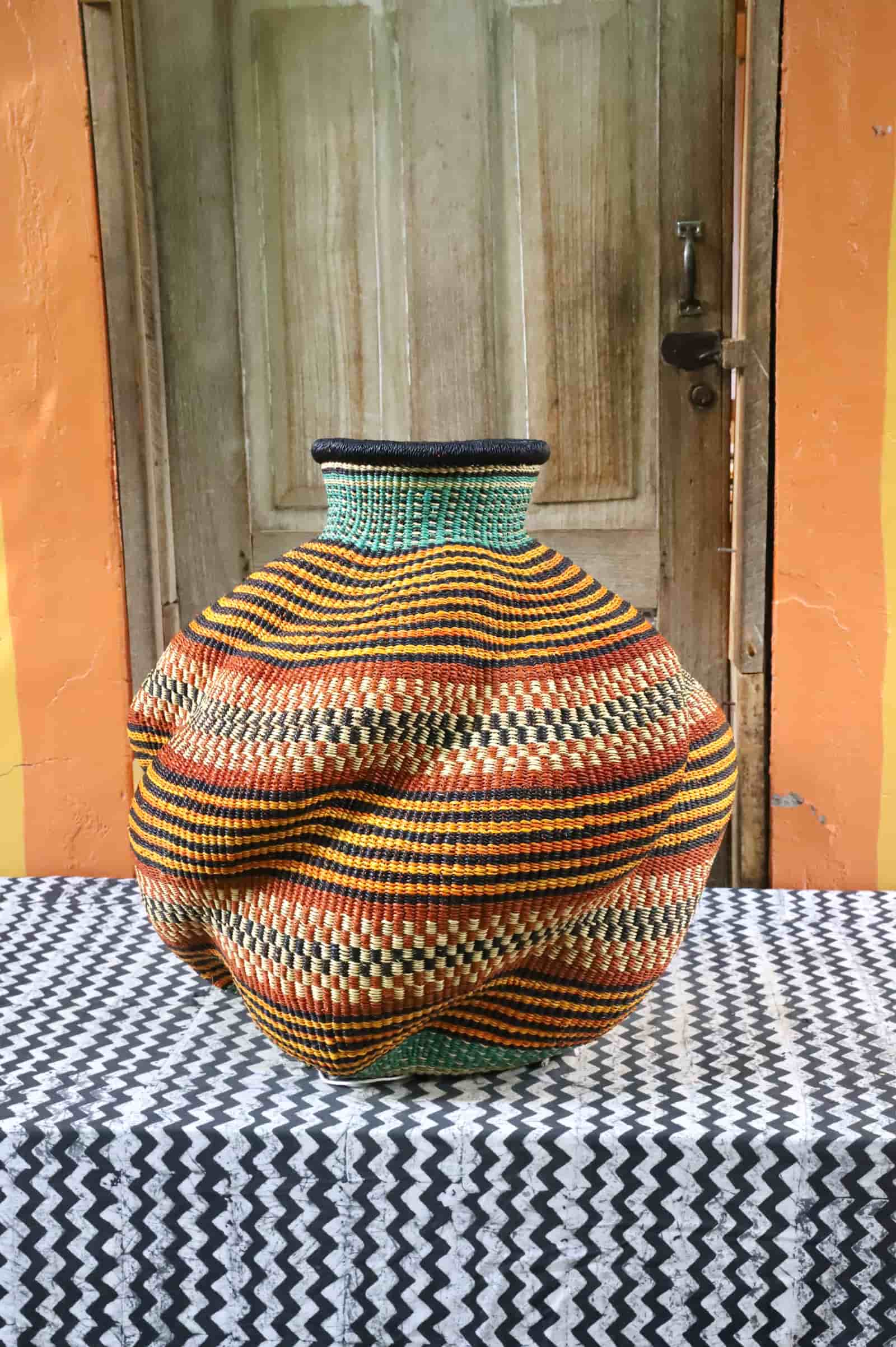 Handwoven De-engo art basket with a lobed gourd form, teal neck and amber, terracotta and black stripes, on a chevron cloth.