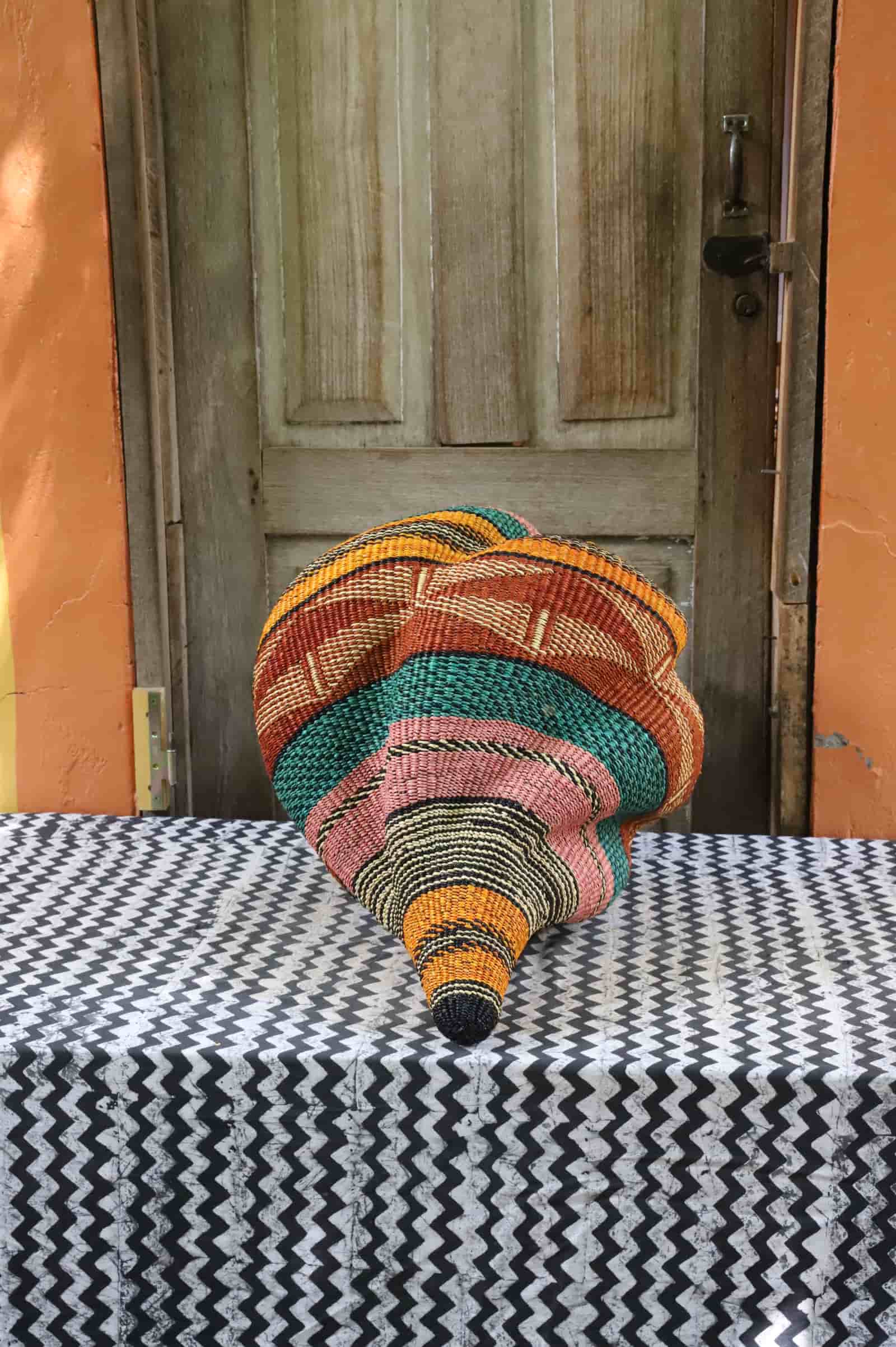 Handwoven Katayeli-tia art basket in teal, terracotta and amber, showing its conch-like spiral form, on a chevron cloth.