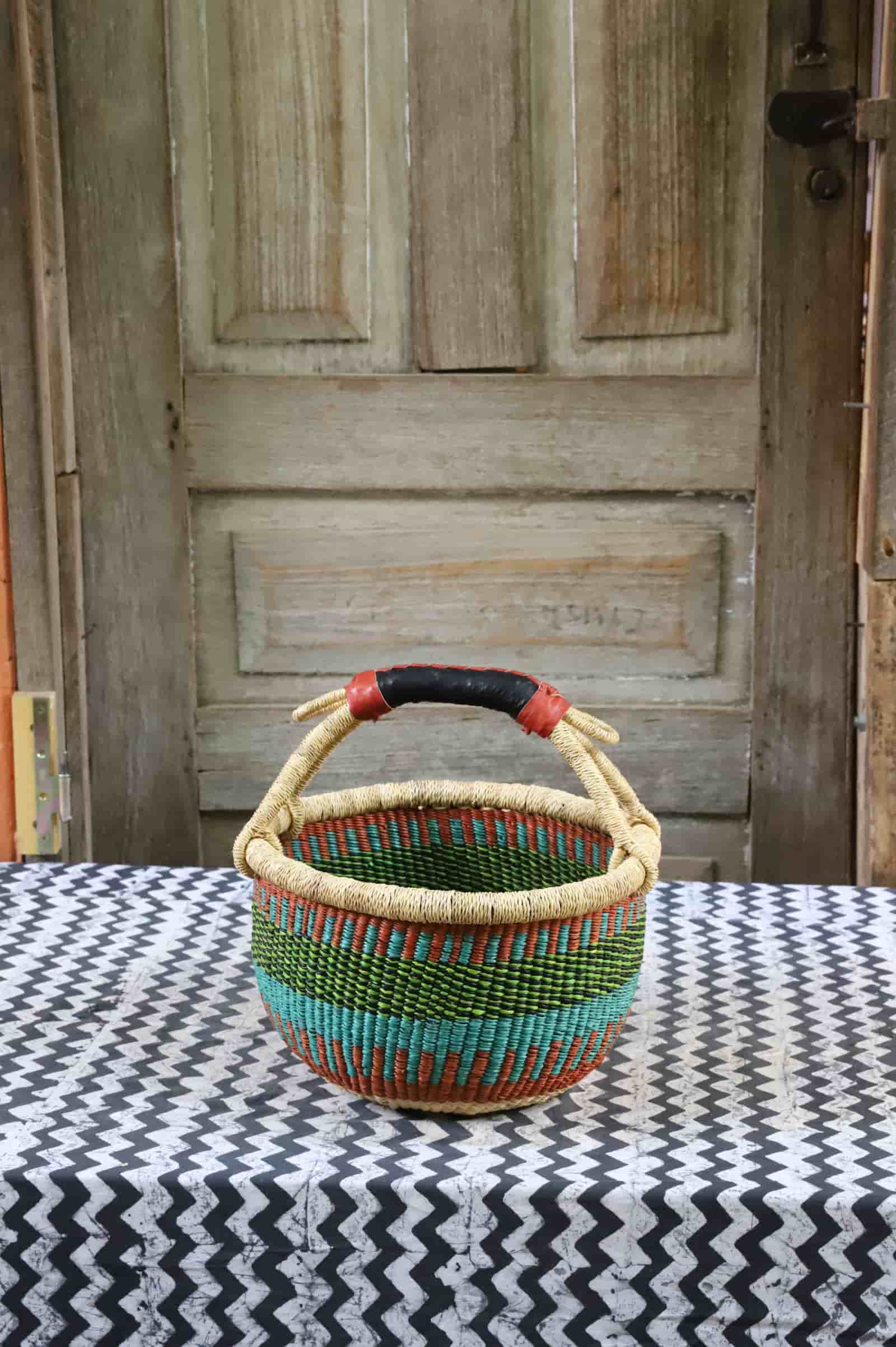 Small round Bolga basket in green, teal and terracotta stripes with a black leather handle, on chevron cloth