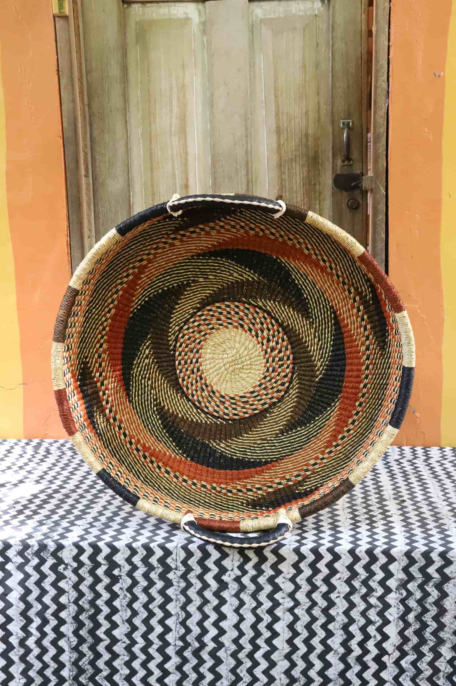 Large double weave woven tray wall décor with dark terracotta and natural swirling pattern, on chevron cloth.
