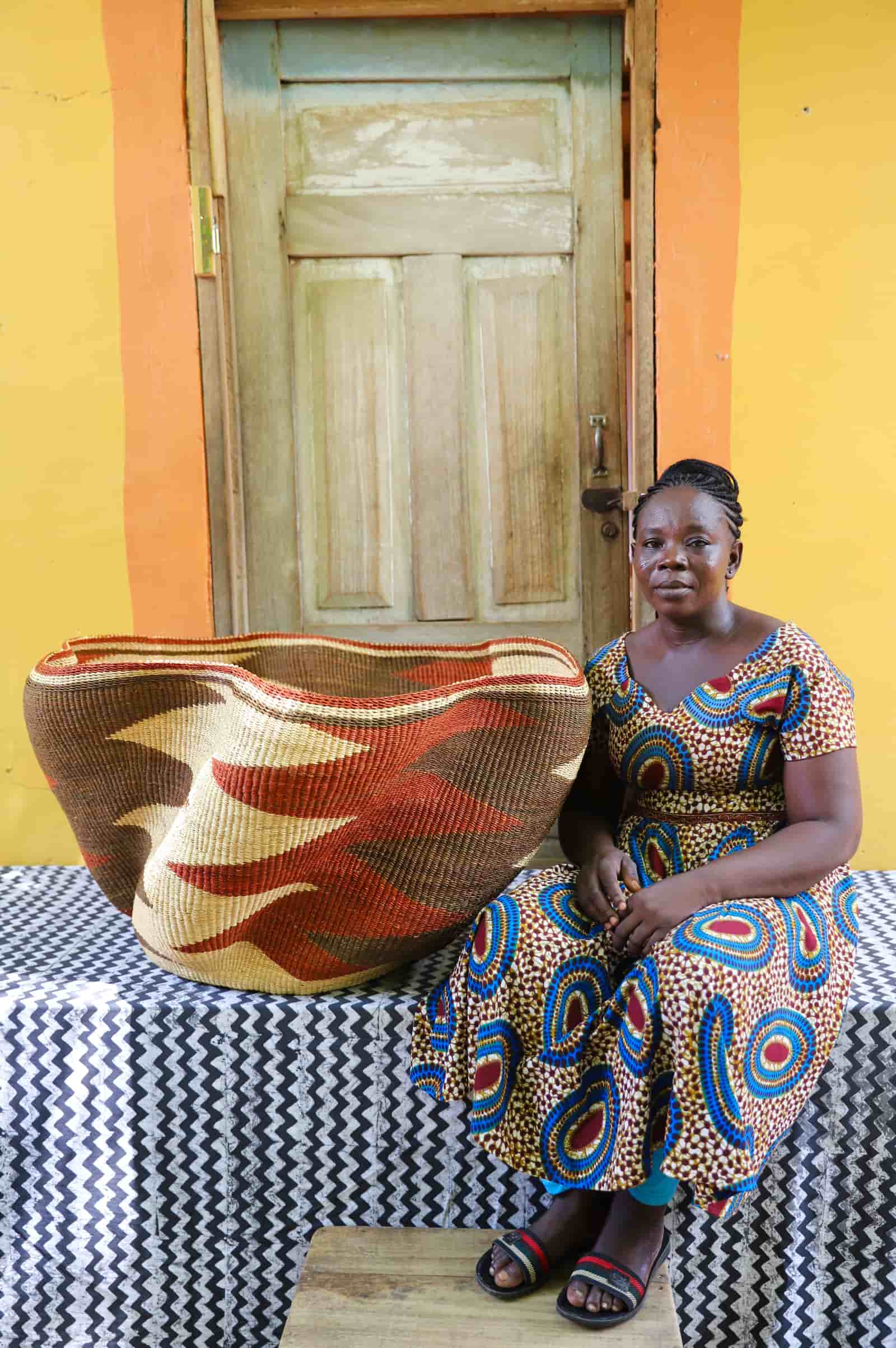 Woman seated beside a Giant Wave art basket with a dark brown and terracotta wave pattern, before a wooden door.