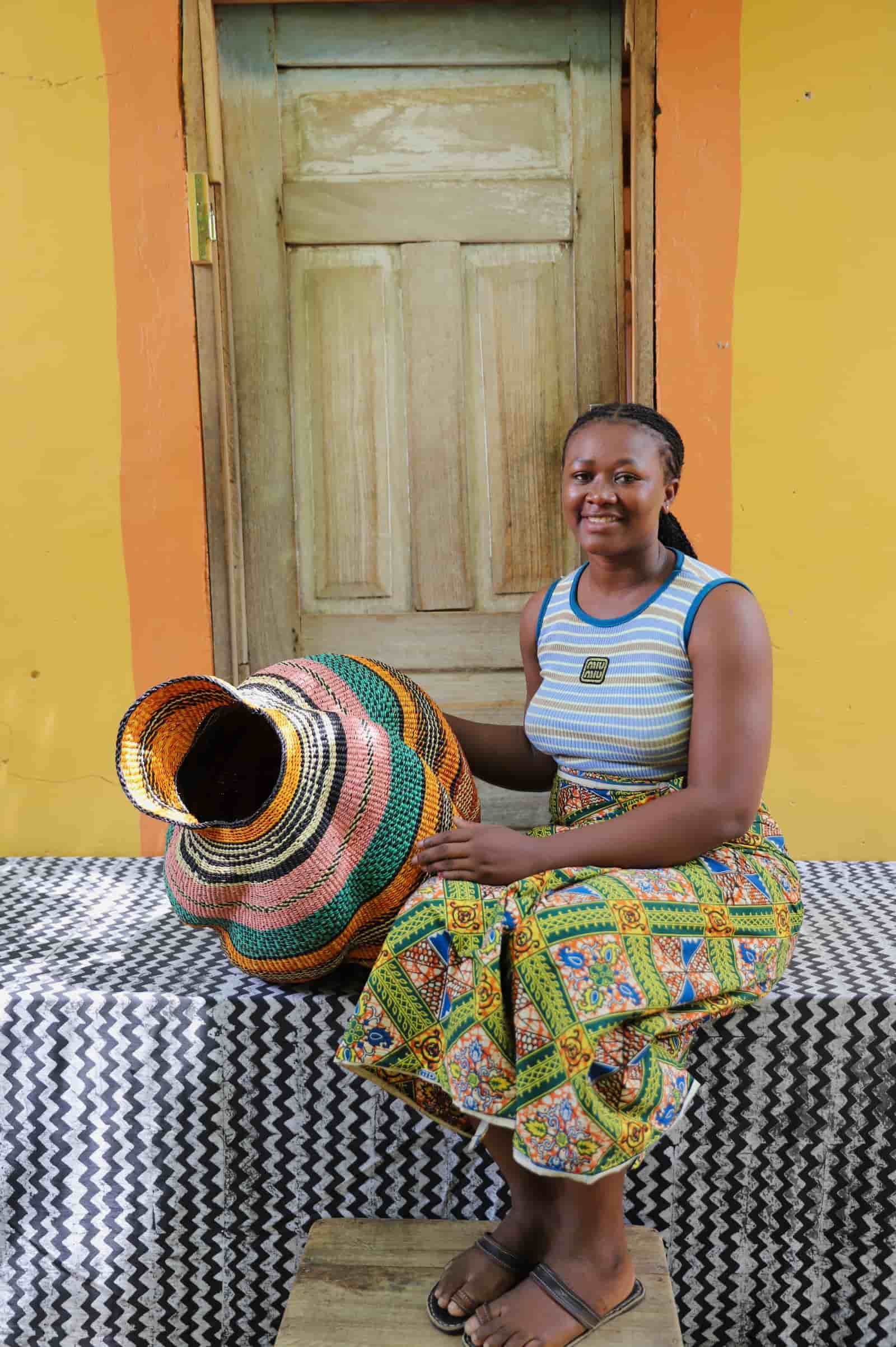 Woman seated beside a handwoven Katayeli-tia art basket in flowing teal, amber and pink stripes, before a wooden door.