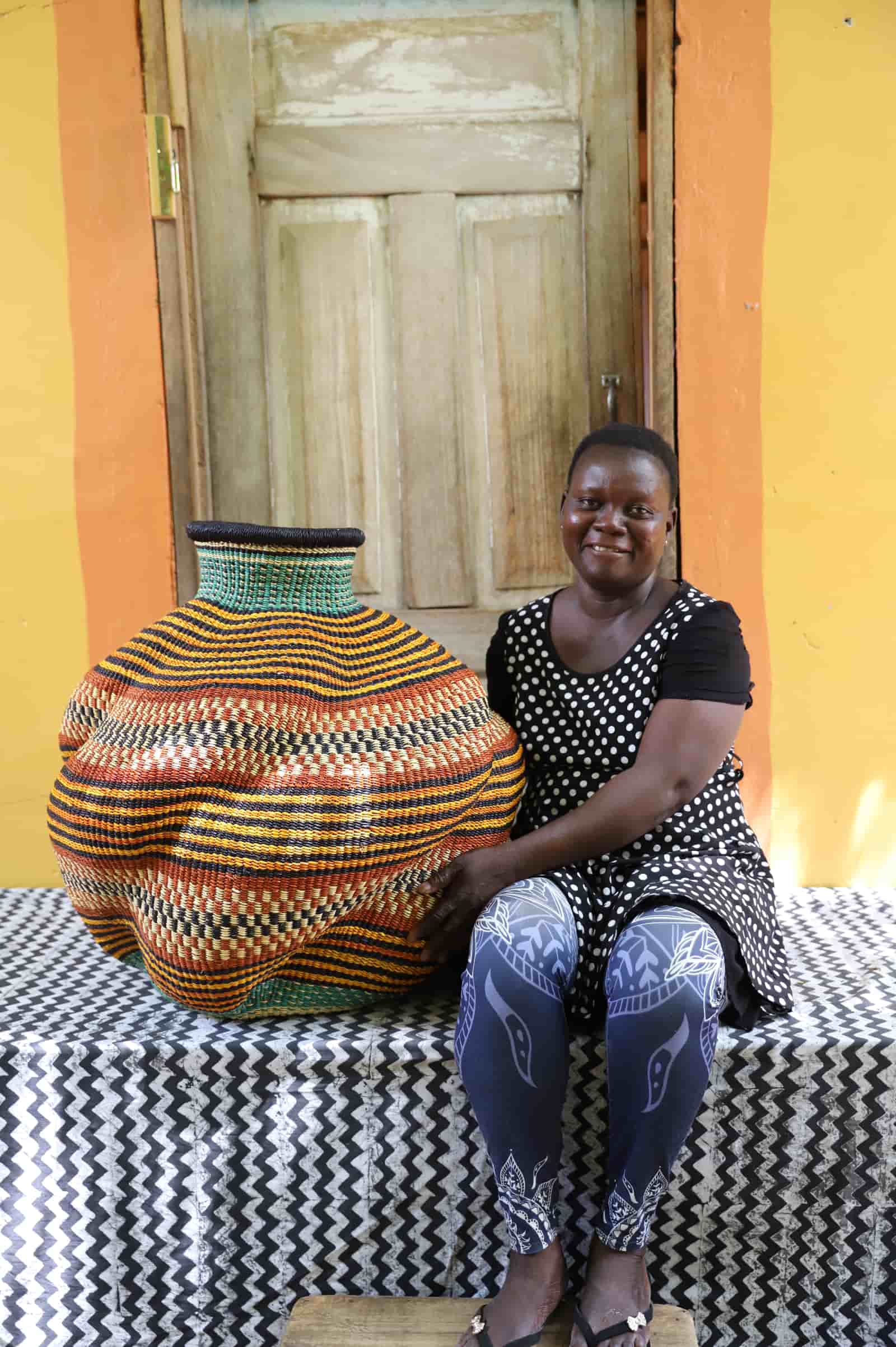 Woman seated beside a handwoven De-engo art basket in amber, terracotta and teal stripes, before a wooden door.