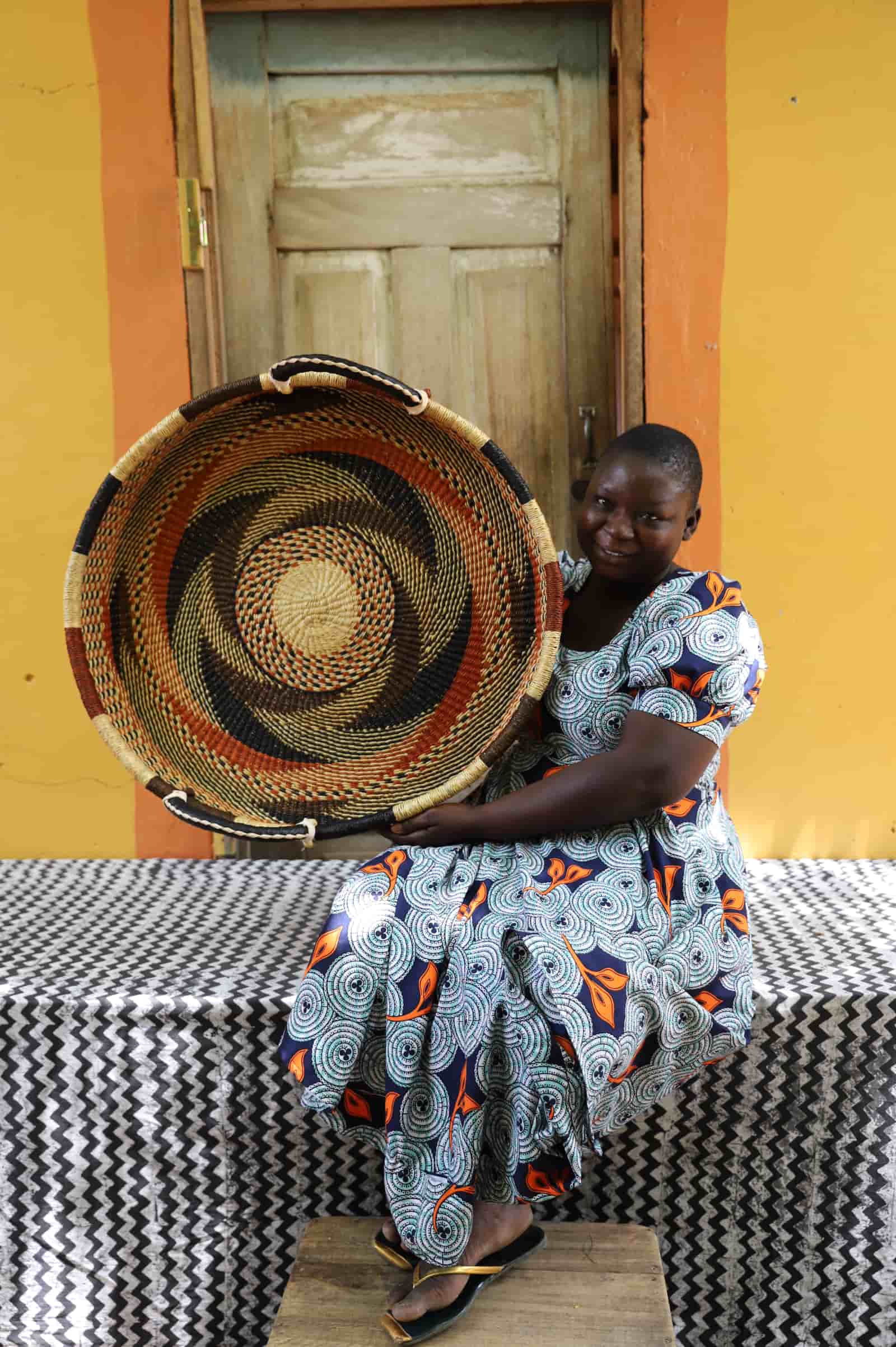 Woman in a patterned dress holding a large double weave woven tray in dark terracotta and natural swirling pattern.