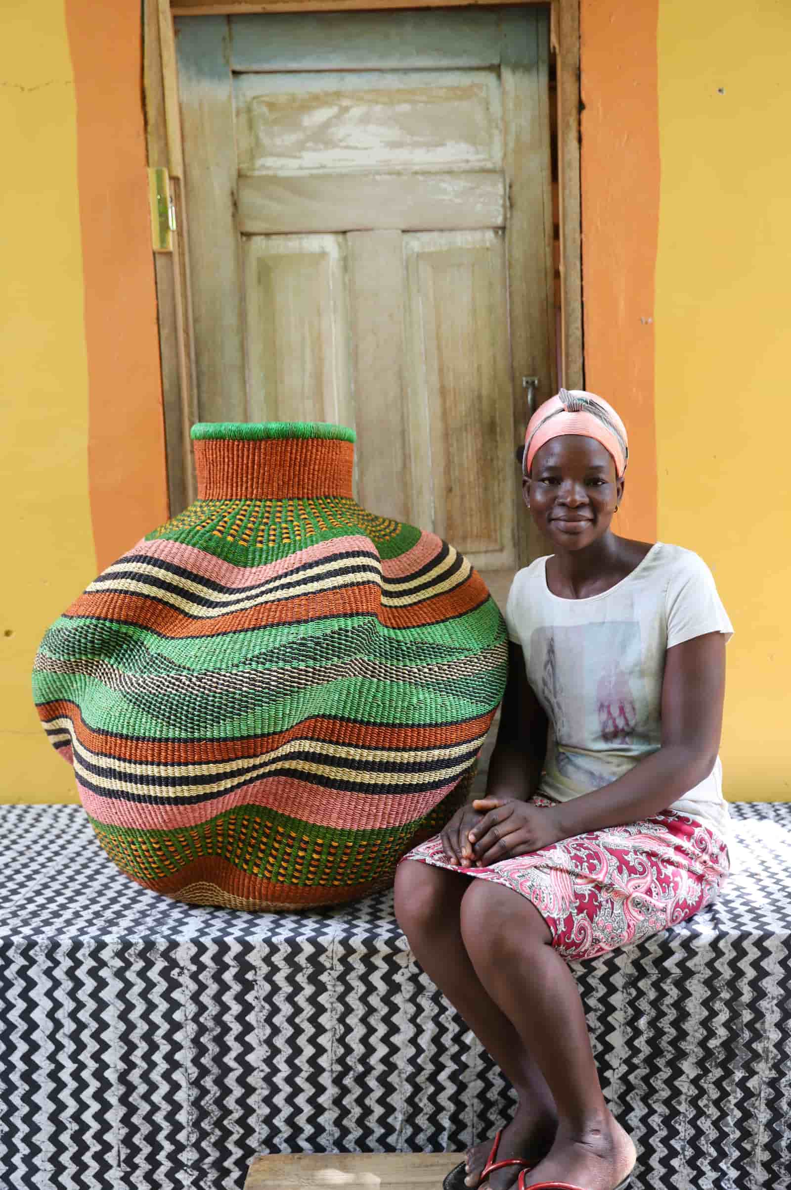 Woman seated beside a large handwoven De-engo art basket in green, terracotta and pink stripes, on a chevron cloth.