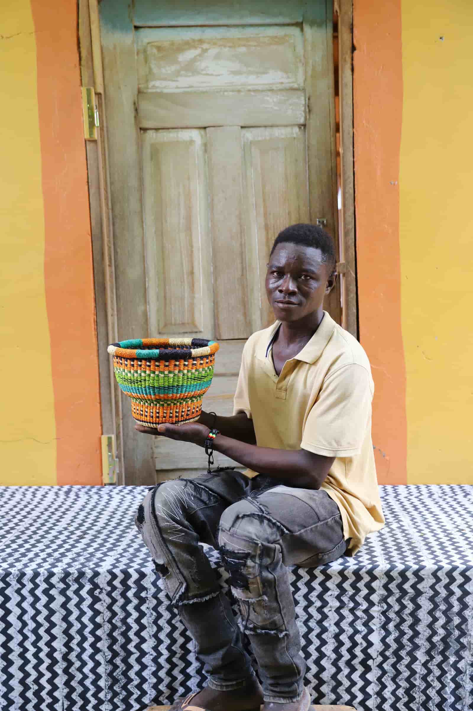 Man holding a tiny handwoven drum storage basket with black, teal, lime and orange stripes, before a wooden door.